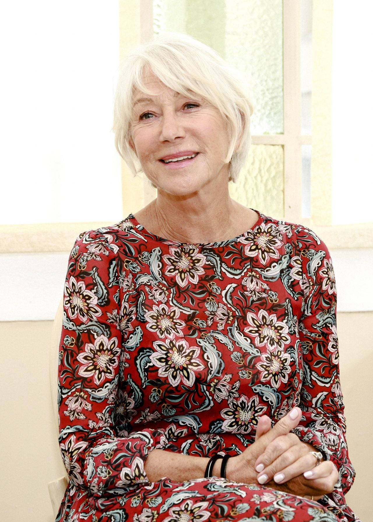 Celebrity 桌布 壁紙 — 一位短白髮的女性微笑著，身穿鮮豔的紅色花卉連衣裙，坐在室內靠近窗邊。