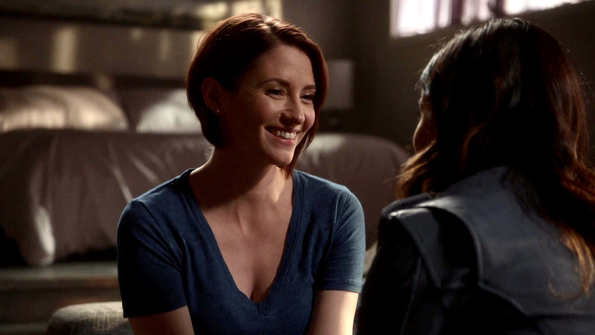 Chyler Leigh 桌布 壁紙 — 一位短紅髮的女性在昏暗的臥室裡坐著，與另一個人交談，露出溫暖的笑容。