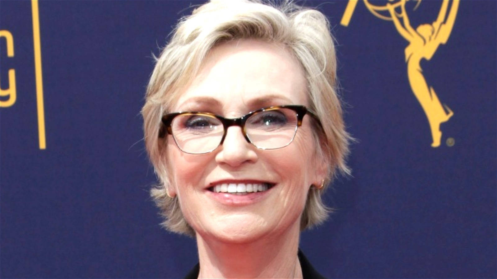 Celebrity 桌布 壁紙 — 一位短金髮、戴玳瑁眼鏡的微笑女性在藍色背景前擺姿勢，背景中有一座金色的艾美獎獎座。