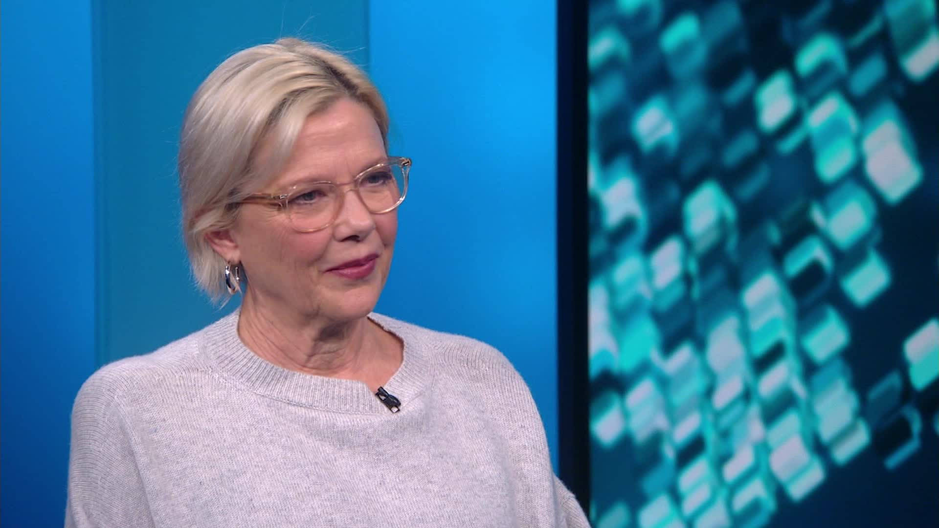 Celebrity 桌布 壁紙 — 一位短金髮、戴眼鏡的女性，穿著淺灰色毛衣，背景是帶有發光幾何圖案的藍色攝影棚。