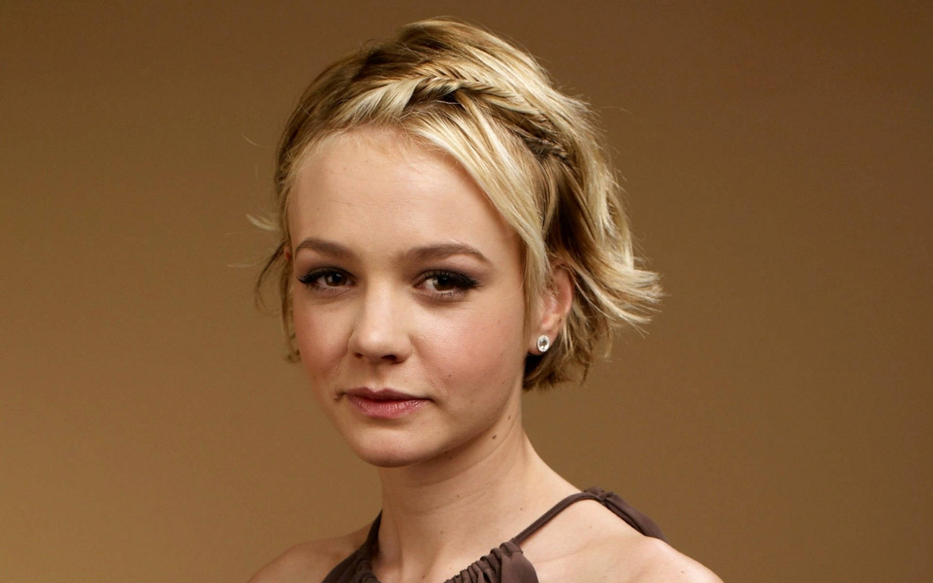 Carey Mulligan 桌布 壁紙 — 一位短金髮、戴辮子頭帶的女性，在溫暖的棕色背景下柔和地凝視著相機。