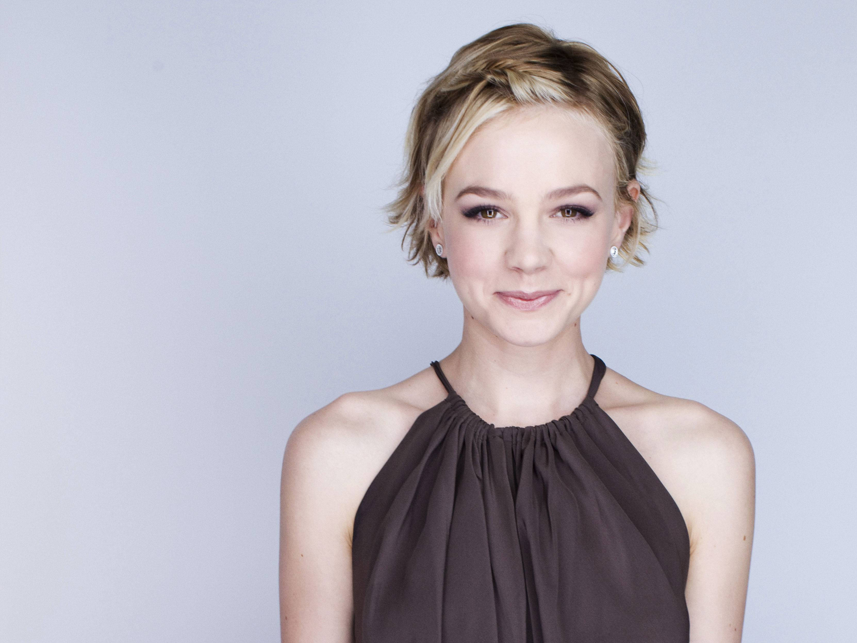 Carey Mulligan 桌布 壁紙 — 一位短金髮、溫柔微笑的女性，在柔和的灰色背景前穿著深色吊帶上衣。