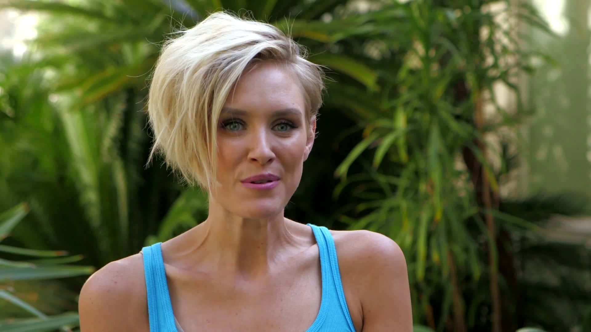 Nicky Whelan 桌布 壁紙 — 一位短金髮、身穿藍色上衣的女性站在茂密的熱帶綠色植物前。
