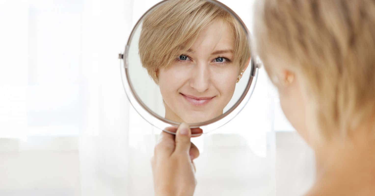 reflection 桌布 壁紙 — 一位短金髮女性在明亮的房間裡拿著圓鏡檢查自己的倒影，溫暖地微笑。
