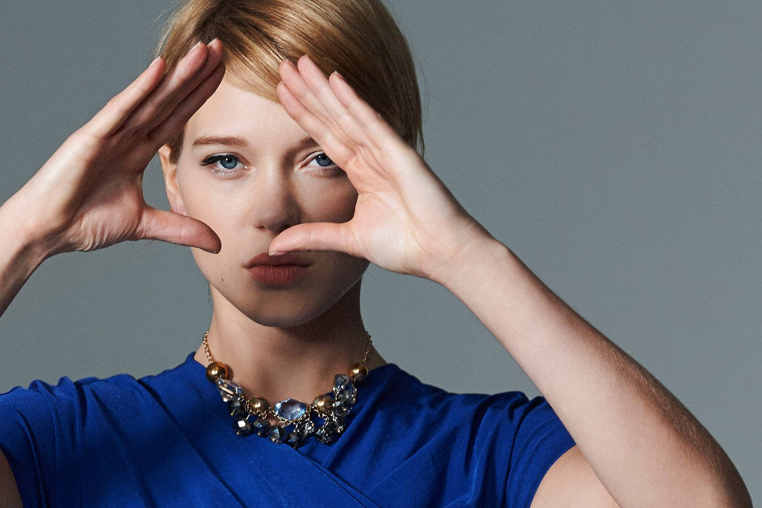 Lea Seydoux 桌布 壁紙 — 一位短金髮女性在灰色攝影棚背景前用手框住臉龐，身穿藍色上衣和珠串項鍊。