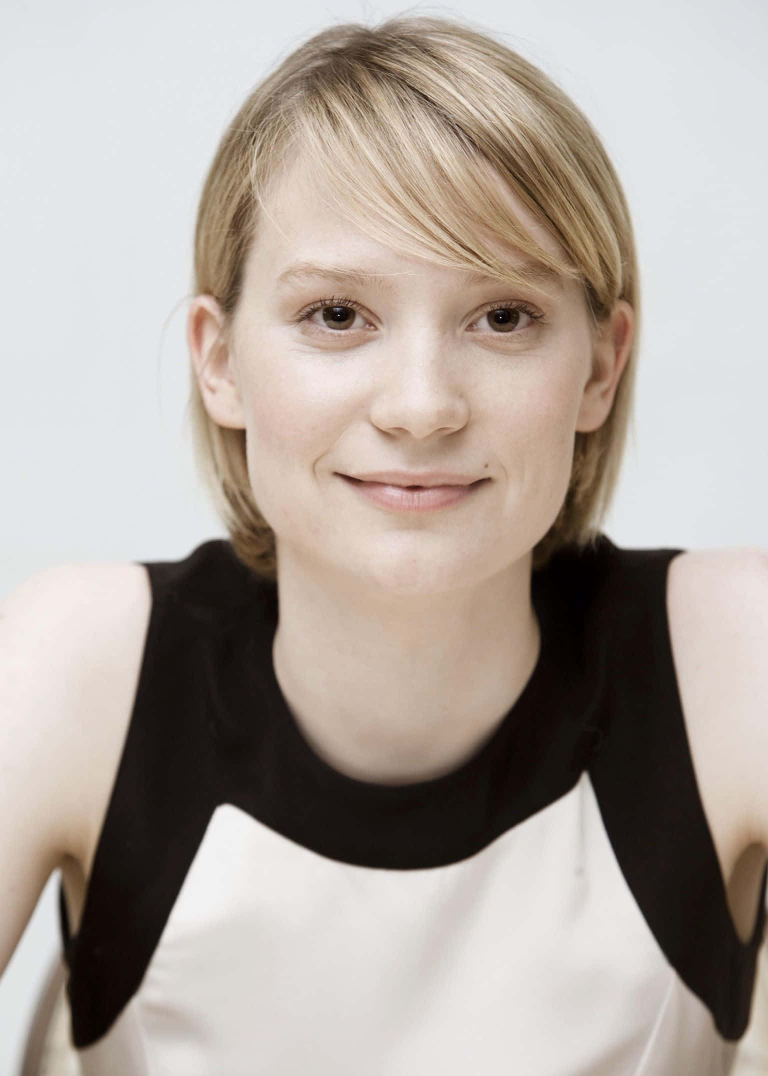 Mia Wasikowska 桌布 壁紙 — 一位短金髮女性在純白背景前微笑，身穿黑白無袖上衣。