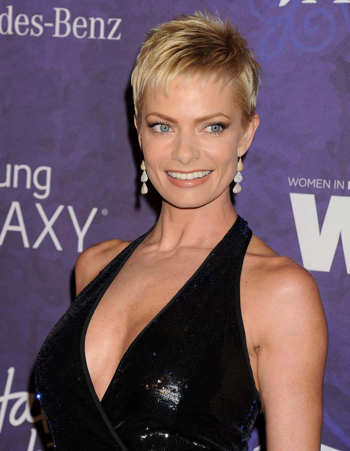 Jaime Pressly 桌布 壁紙 — 一位短金髮女性在紫色品牌活動背景前穿著黑色亮片連衣裙微笑。