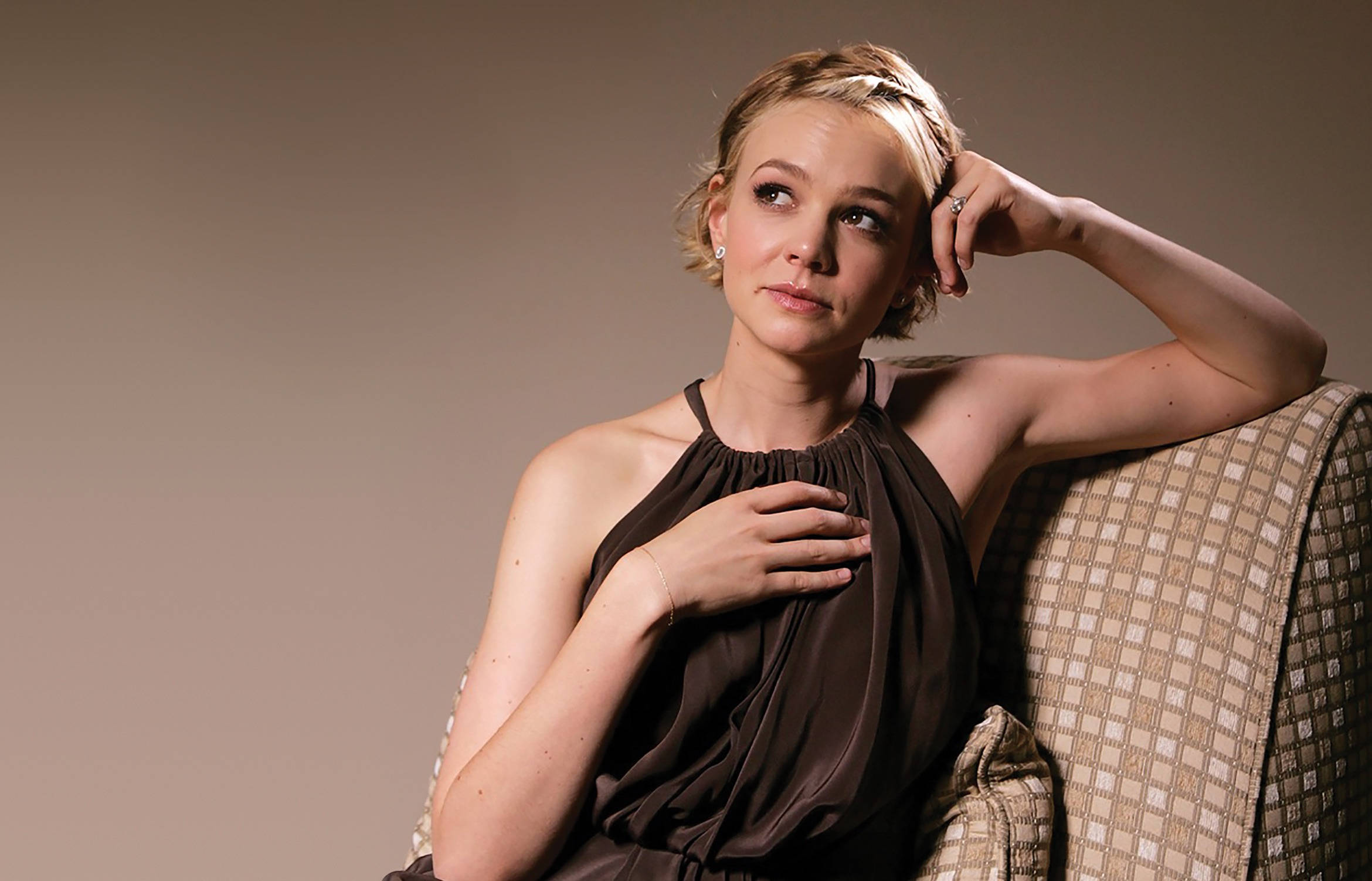 Carey Mulligan 桌布 壁紙 — 一位短金髮女性坐在圖案沙發上，手放在胸前，沉思地向上看，表情平靜。
