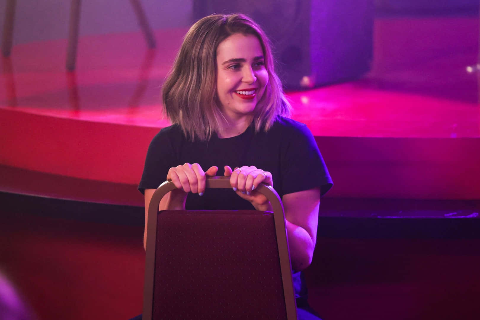 Mae Whitman 桌布 壁紙 — 一位短金髮微笑的女性在鮮豔的粉紅和紫色舞台燈光下抓住椅背。