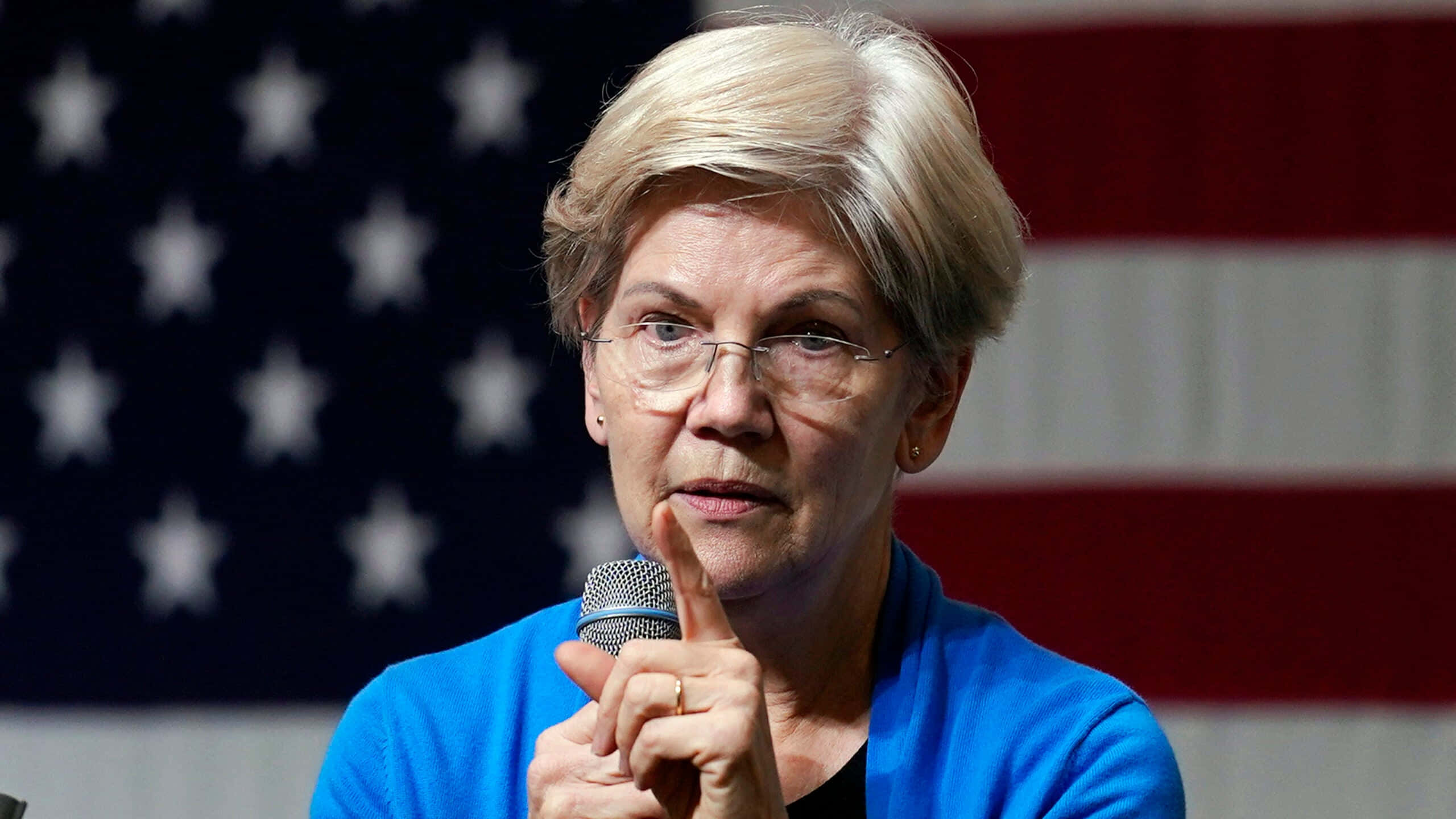 Politics 桌布 壁紙 — 一位短金髮戴眼鏡的女性，在美國國旗背景下，對著麥克風說話，同時手指向前方。