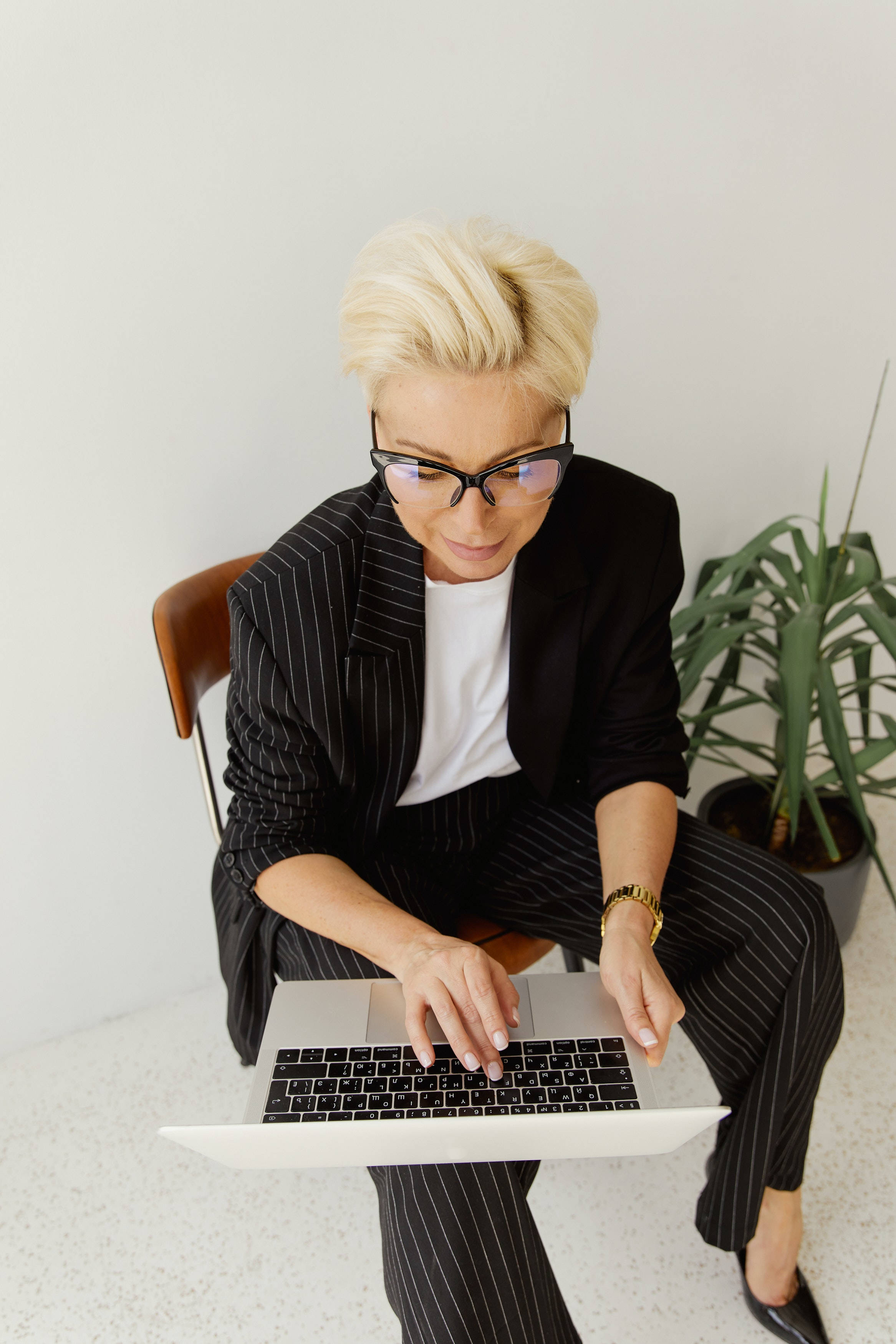aesthetic tumblr laptop 桌布 壁紙 — 一位短金髮戴眼鏡的女性坐在簡約風格的房間裡，旁邊有一盆盆栽，正在使用筆記型電腦打字。