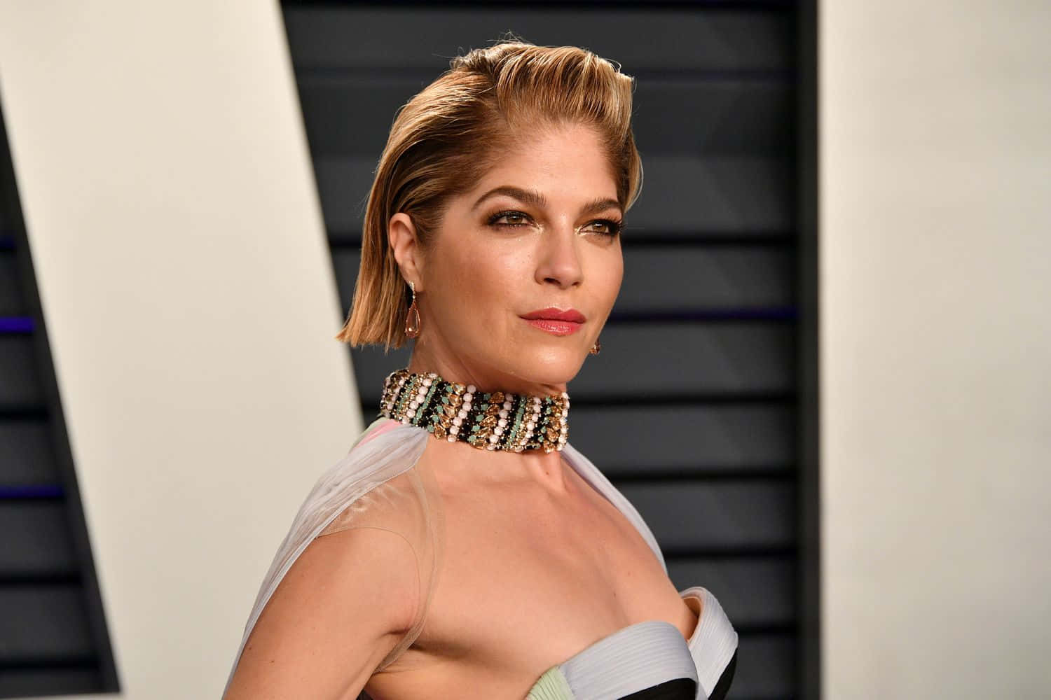 Selma Blair 桌布 壁紙 — 一位短金髮的優雅女士，戴著珠串頸鏈，身穿透視長袍，站在現代幾何背景前。