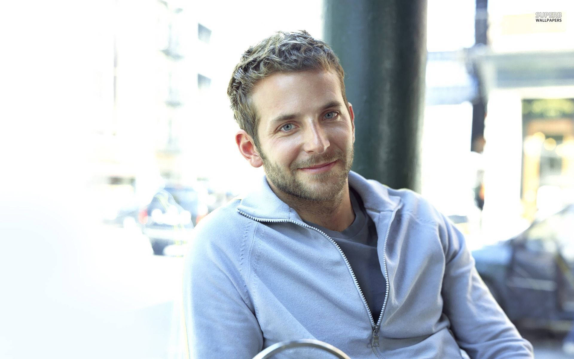 Bradley Cooper 桌布 壁紙 — 一位短髮、留著淺鬍鬚的男子，穿著淺灰色拉鍊連帽衫，溫暖地微笑。