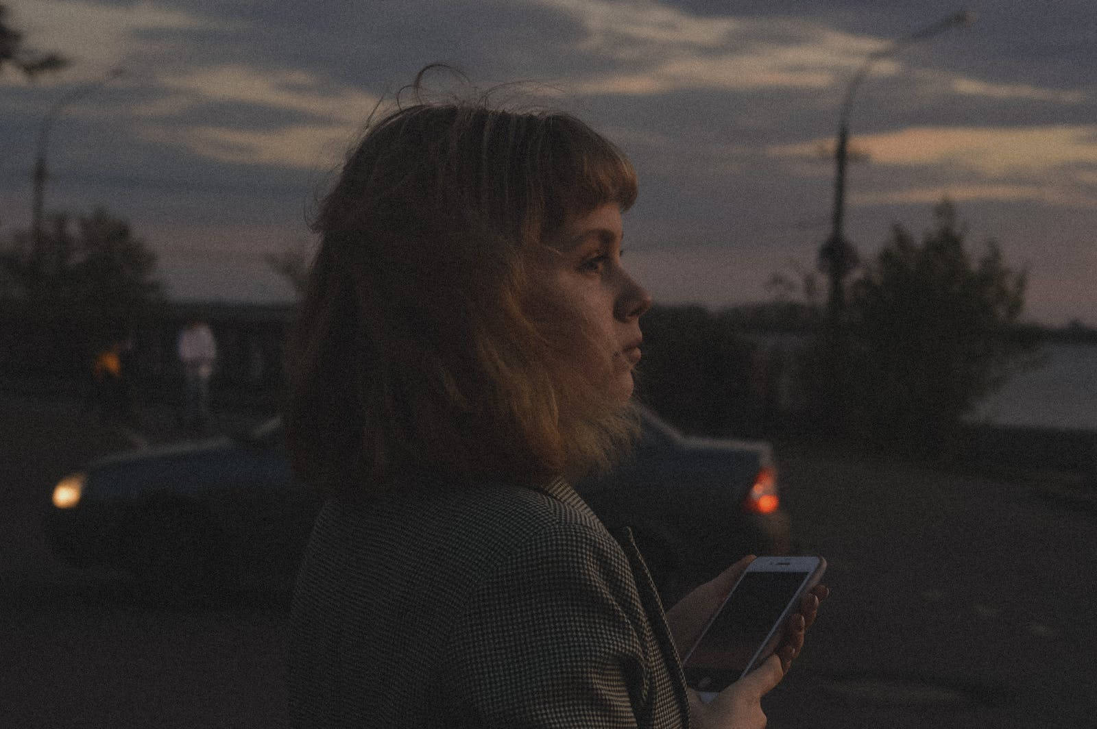 深色 iPhone 桌布 壁紙 — 一位短髮女性在黃昏時分站在寧靜的街道上，手持電話，身後有汽車駛過。