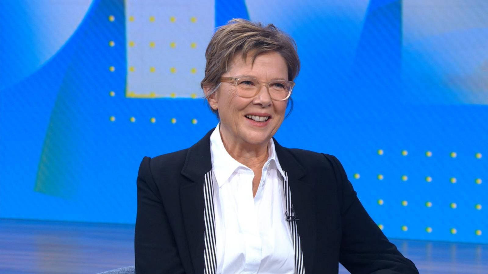 Celebrity 桌布 壁紙 — 一位短髮戴眼鏡的 smiling 女性，在藍色幾何背景前穿著黑色西裝外套內搭白色條紋襯衫。