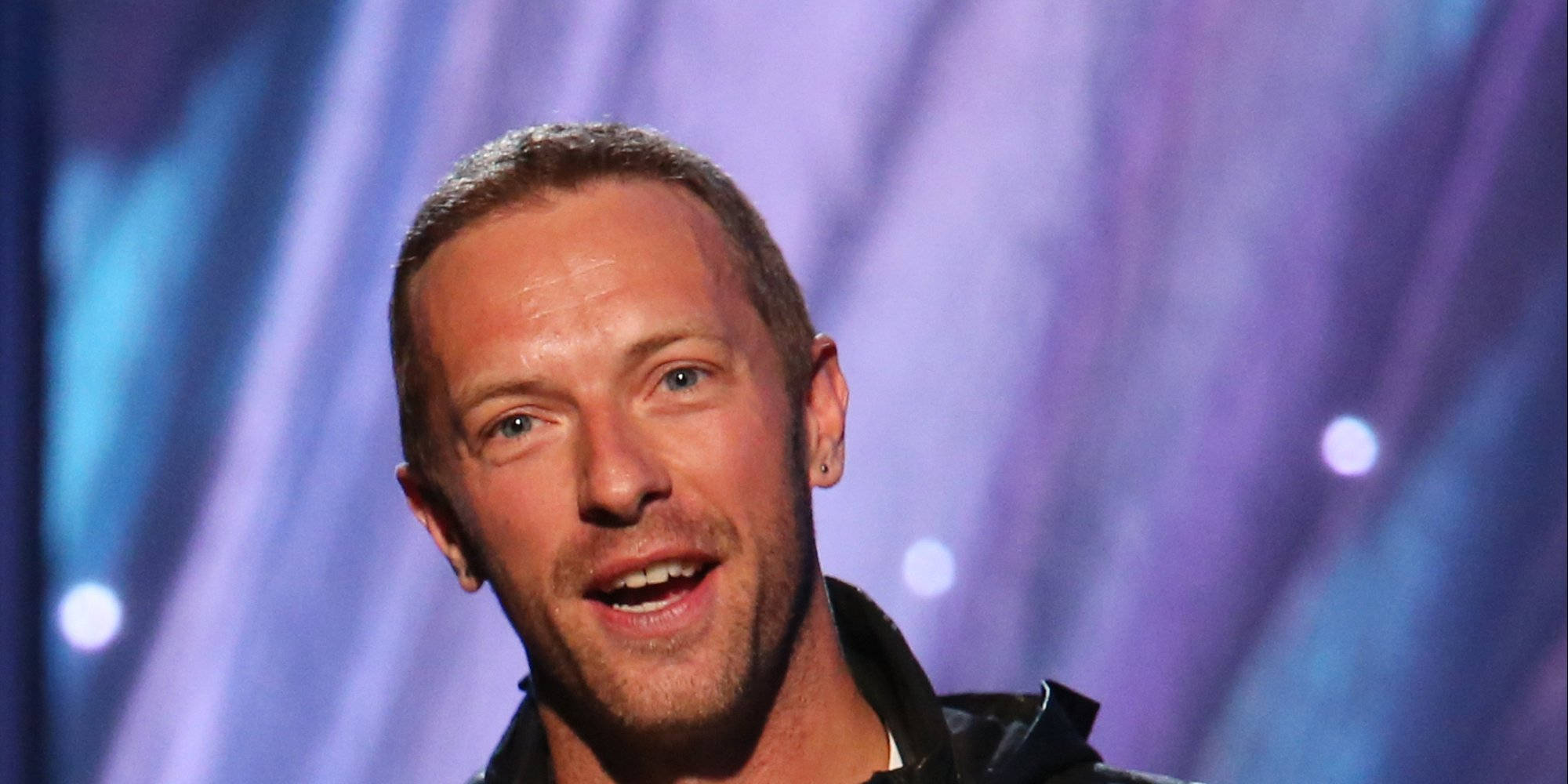 Chris Martin 桌布 壁紙 — 一位短髮藍眼的男士在充滿活力的紫色和藍色舞台背景及發光燈光下溫暖地微笑。