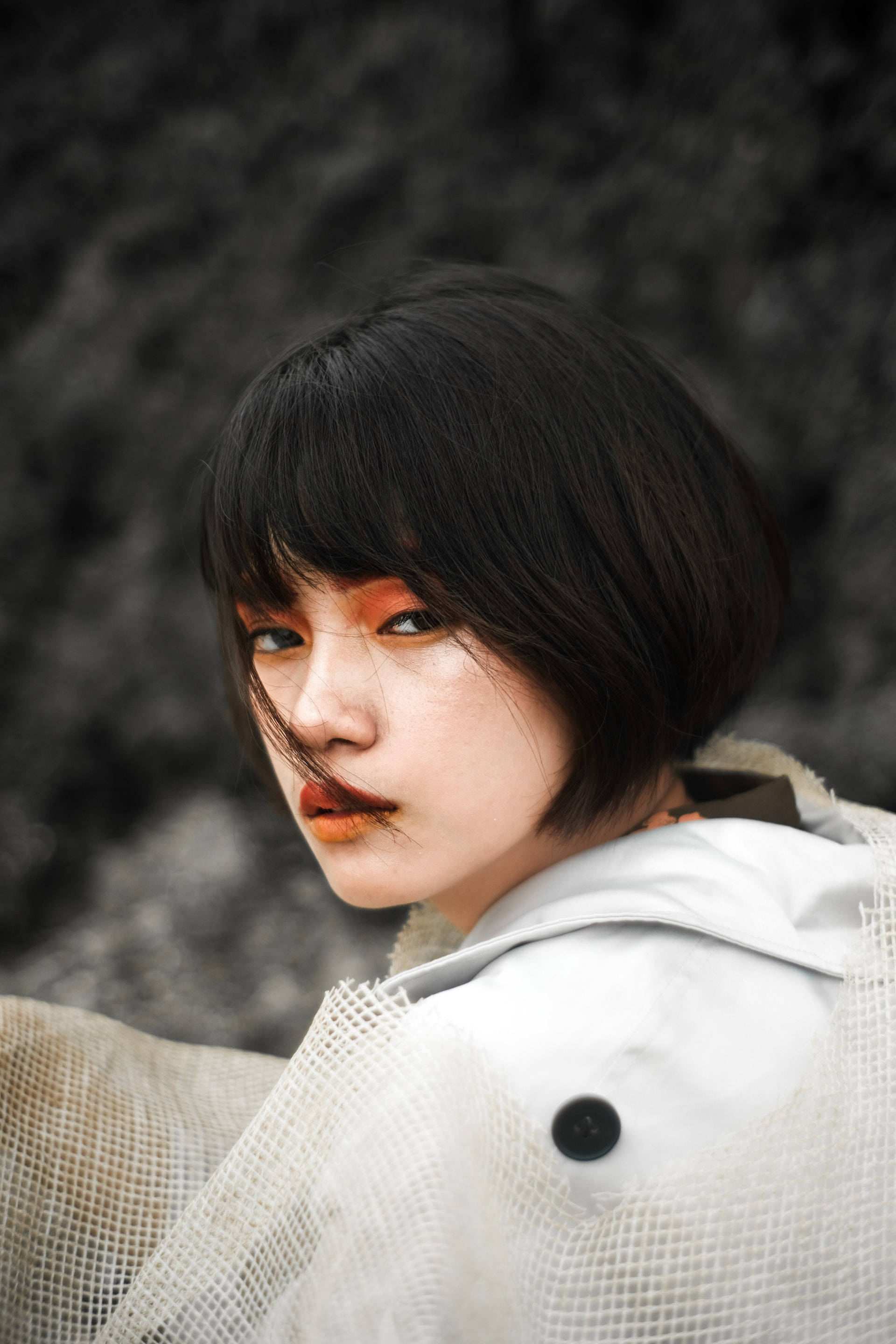 lovely girl 桌布 壁紙 — 一位短黑髮、化著大膽橙色妝容的女性，在模糊的灰色背景下，穿著質感白色外套，目光強烈地凝視著。