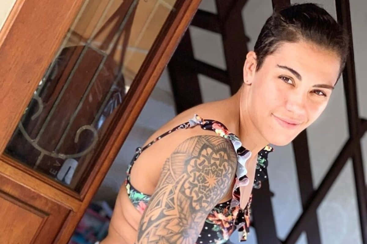 MMA 桌布 壁紙 — 一位短黑髮、左臂上有大型幾何刺青的女士靠在木門框上微笑。