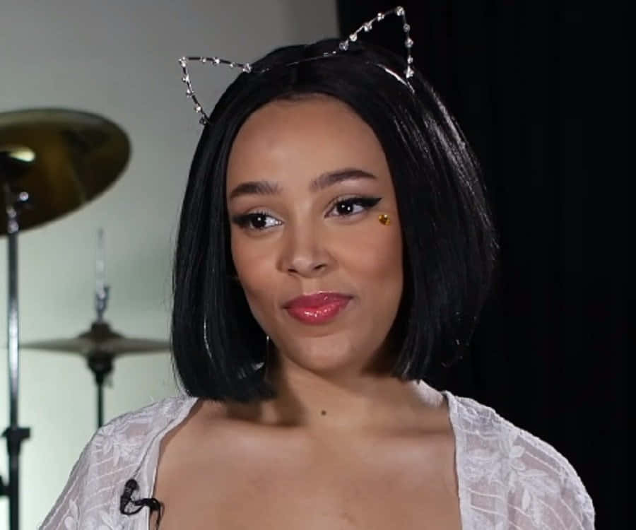 Doja Cat 桌布 壁紙 — 一位短黑髮、戴貓耳的女性，穿著白色蕾絲上衣，身後有爵士鼓，溫柔地微笑。