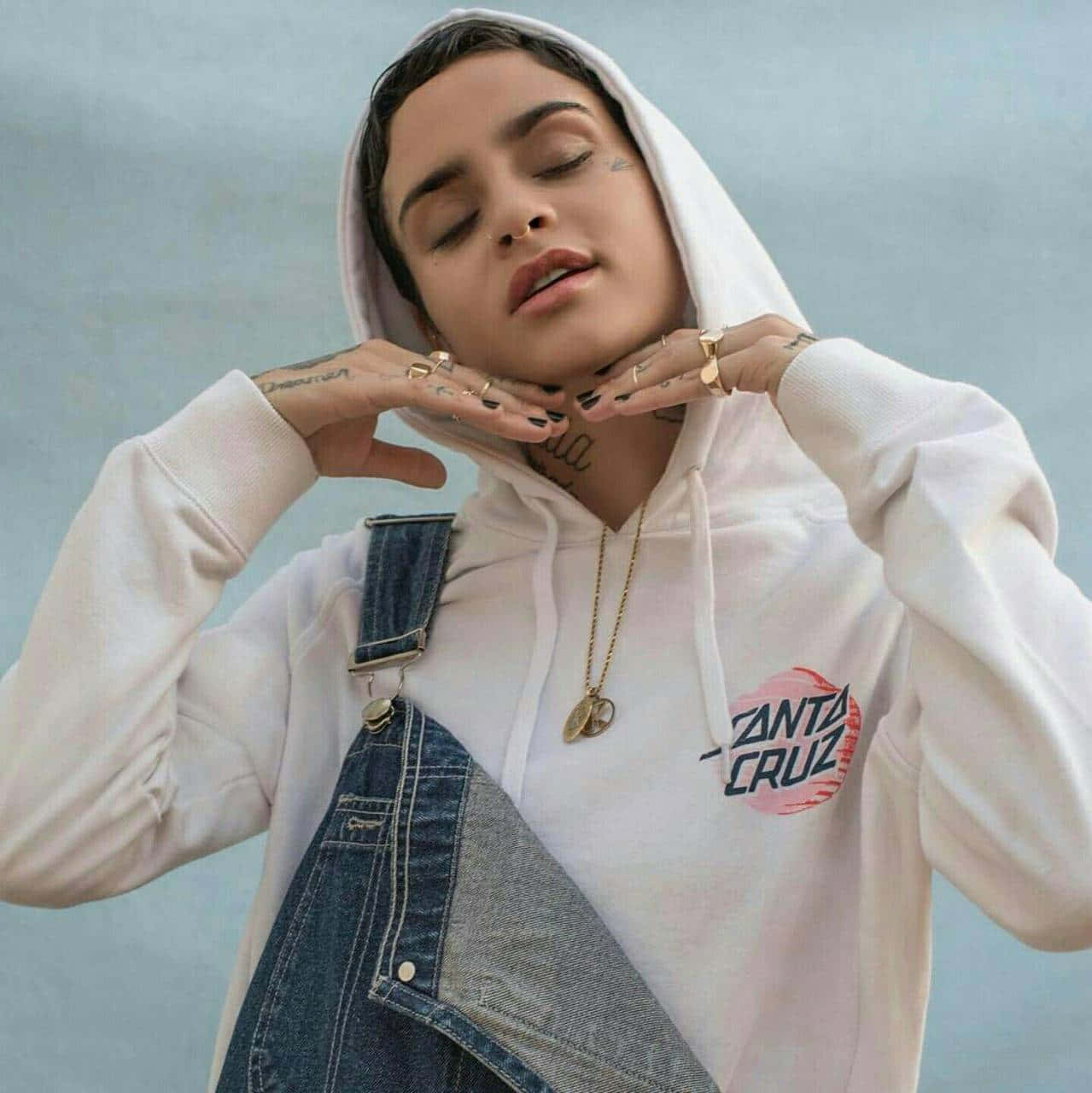 kehlani 桌布 壁紙 — 一位短黑髮、有刺青的年輕女性，穿著白色連帽衫和牛仔吊帶褲，在柔和的藍色背景前將手放在脖子附近擺姿勢。