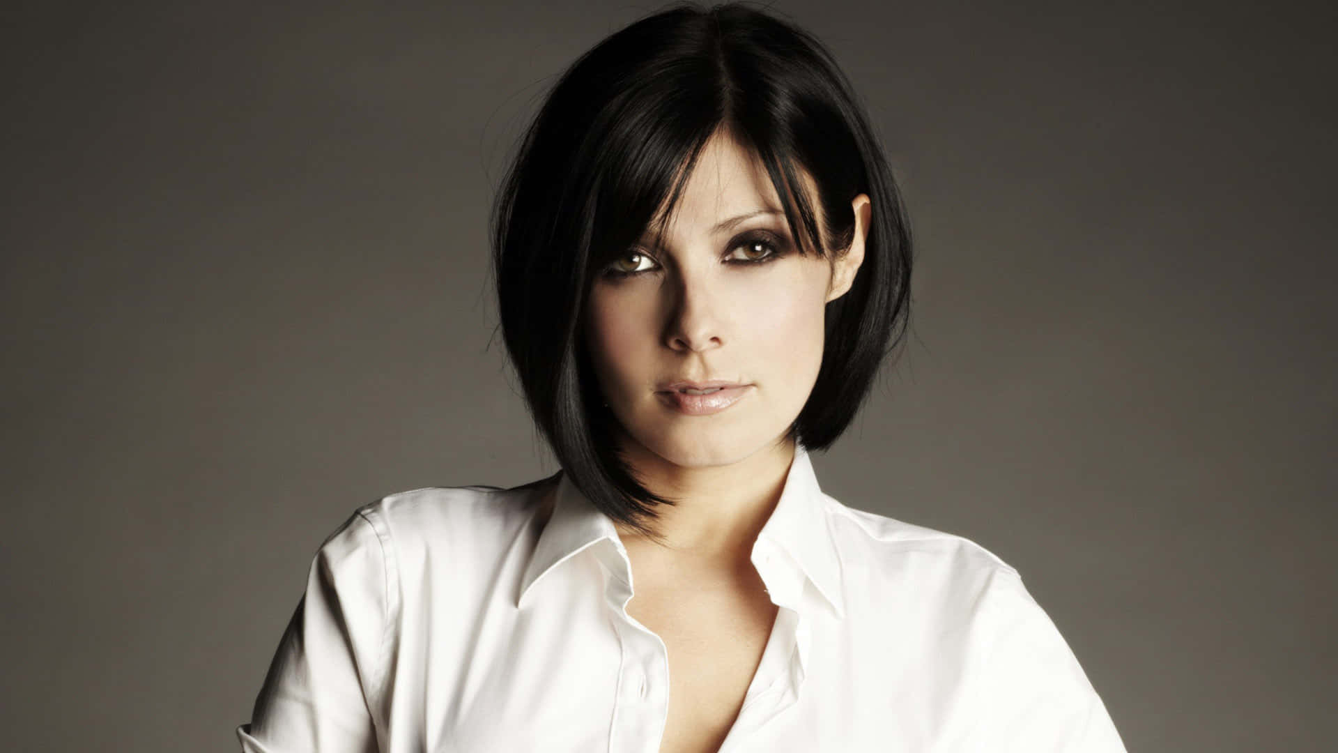 Kym Marsh 桌布 壁紙 — 一位短黑髮、煙燻妝眼睛的女性，穿著一件清爽的白色鈕扣襯衫，在柔和的灰色背景前直視鏡頭。