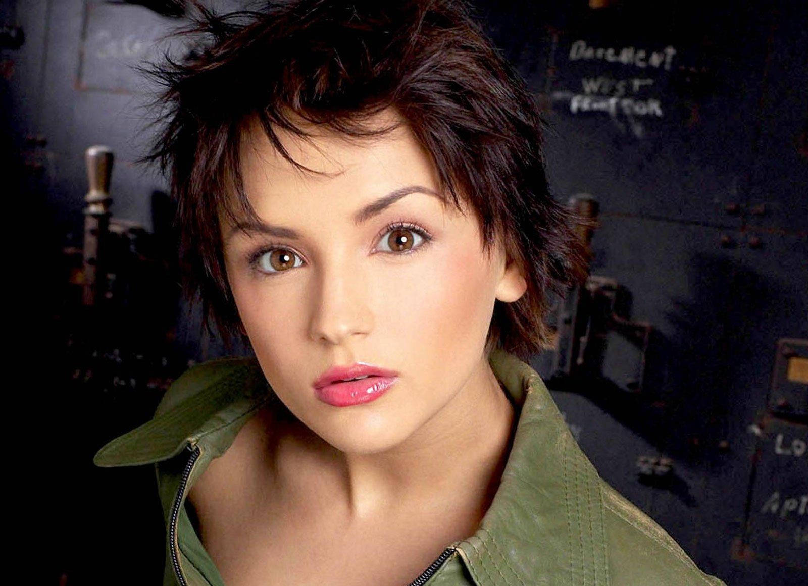 Actress HD 桌布 壁紙 — 一位短黑髮、粉紅嘴唇的女性，穿著綠色外套，在黑暗的工業背景下直視鏡頭。