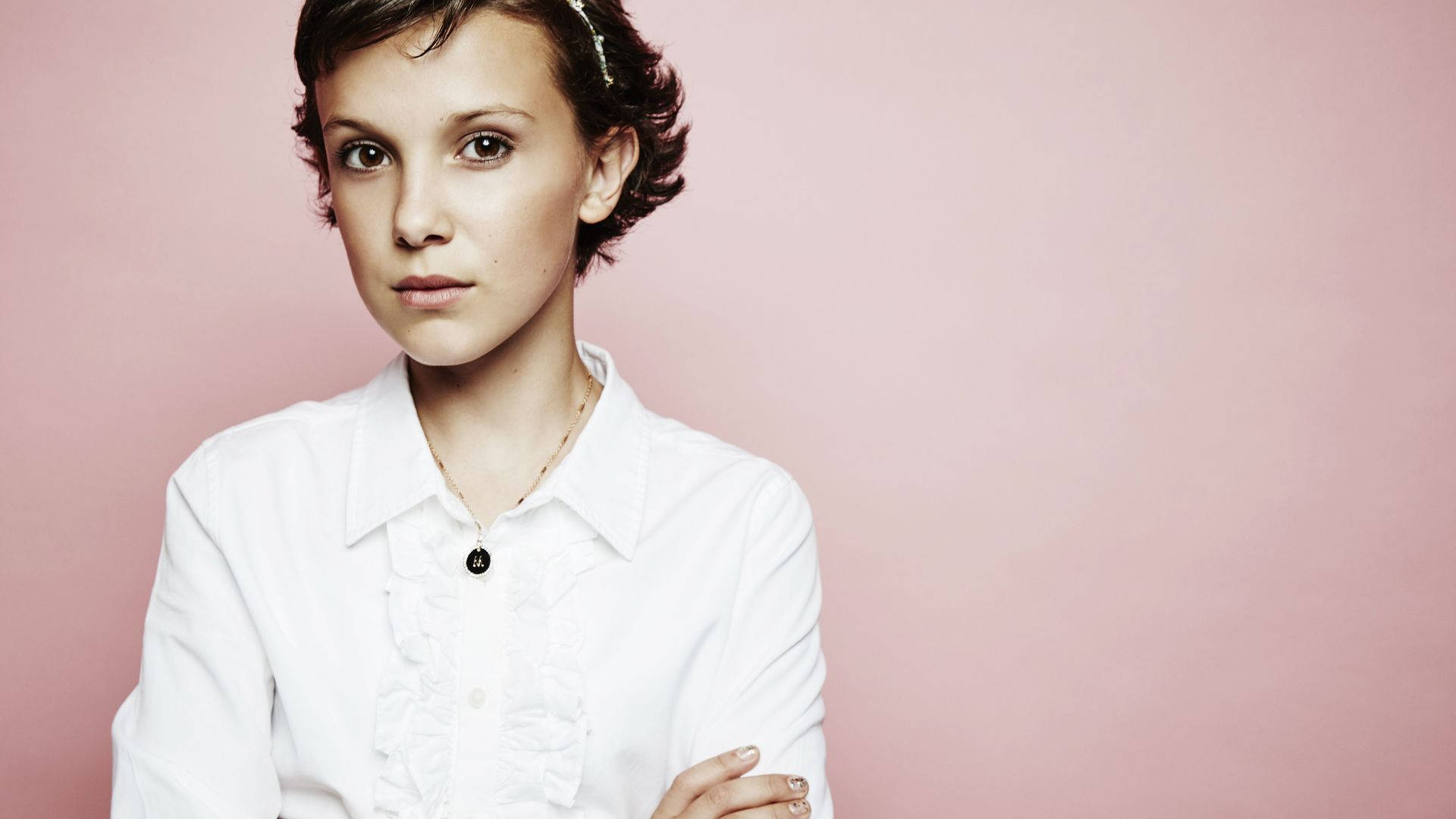 Millie 桌布 壁紙 — 一位短黑髮、身穿白色襯衫的年輕女性，雙臂交叉，站在柔和的粉紅色背景前。