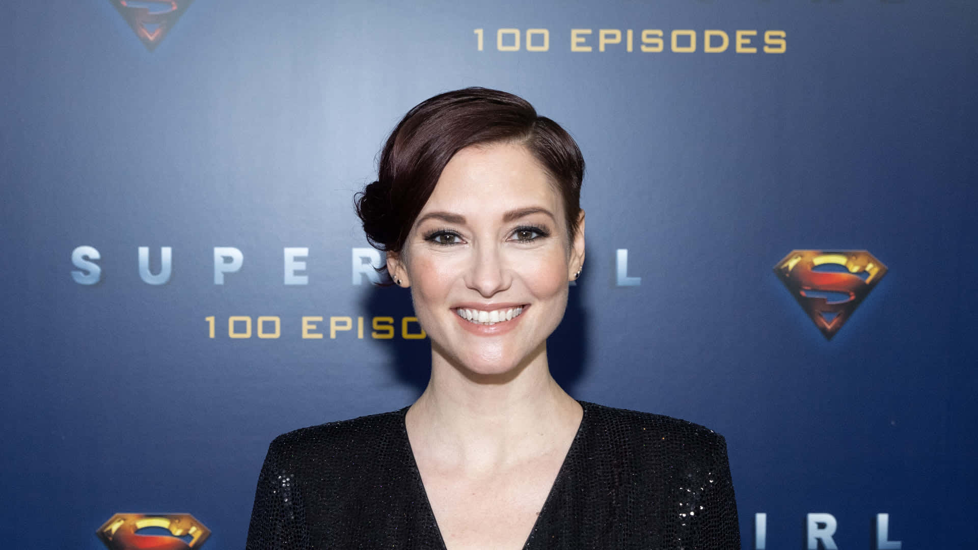Chyler Leigh 桌布 壁紙 — 一位短黑髮、身穿黑色亮片上衣的微笑女士站在藍色「Supergirl 100 Episodes」活動背景前。
