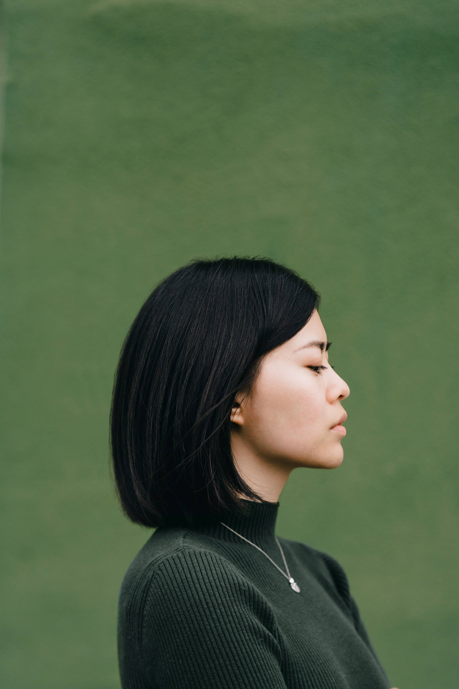 Woman 桌布 壁紙 — 一位短黑髮女性側身站在柔和的綠色牆壁前，閉著眼睛靜靜沉思。