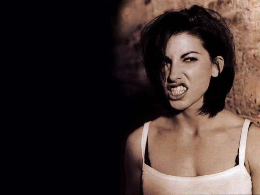 Gina Gershon 桌布 壁紙 — 一位短黑髮女性在戲劇性的低調燈光下，對著有紋理的牆壁做出兇狠的鬼臉。