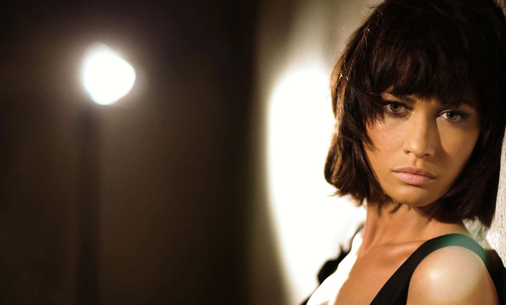 Olga Kurylenko 桌布 壁紙 — 一位短黑髮女性在戲劇性的工作室燈光下，背景有明亮的聚光燈，強烈地凝視著相機。