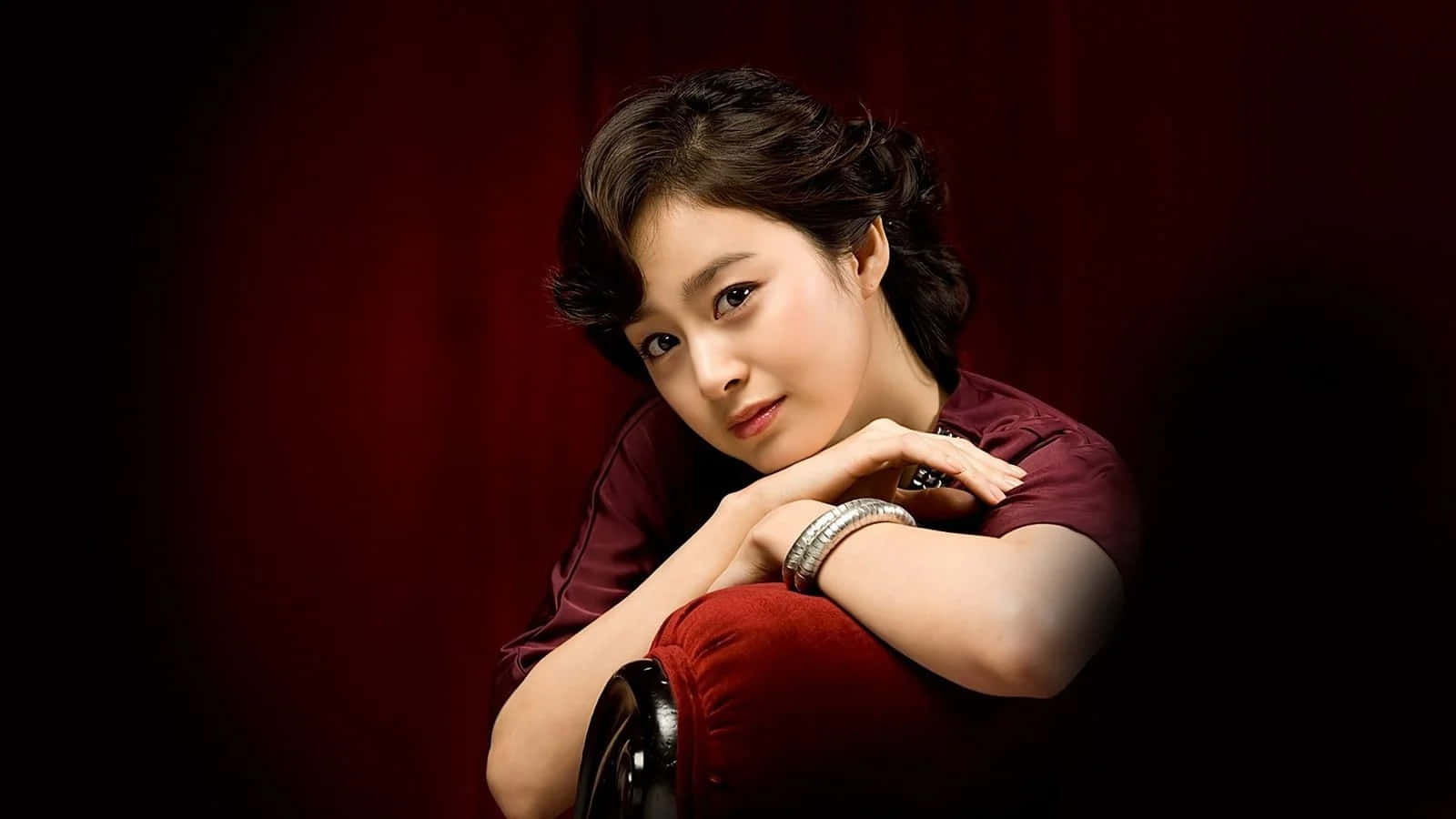 女演員 桌布 壁紙 — 一位短黑髮女性將下巴擱在手上，背景是深紅色天鵝絨，身穿相配的栗色上衣和銀色手鐲。