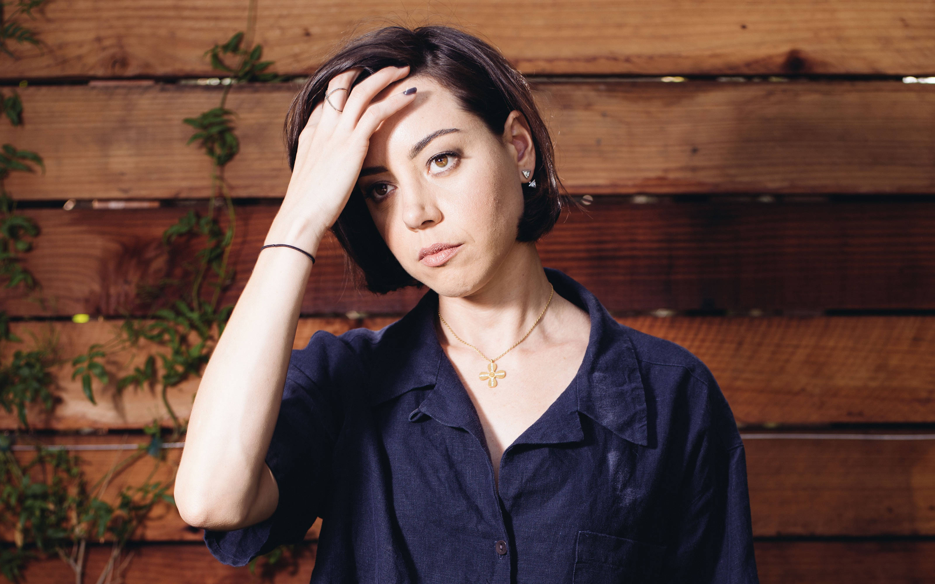 Aubrey 桌布 壁紙 — 一位短黑髮女性將手放在額頭上，靠著一面有綠色藤蔓的質樸木牆。