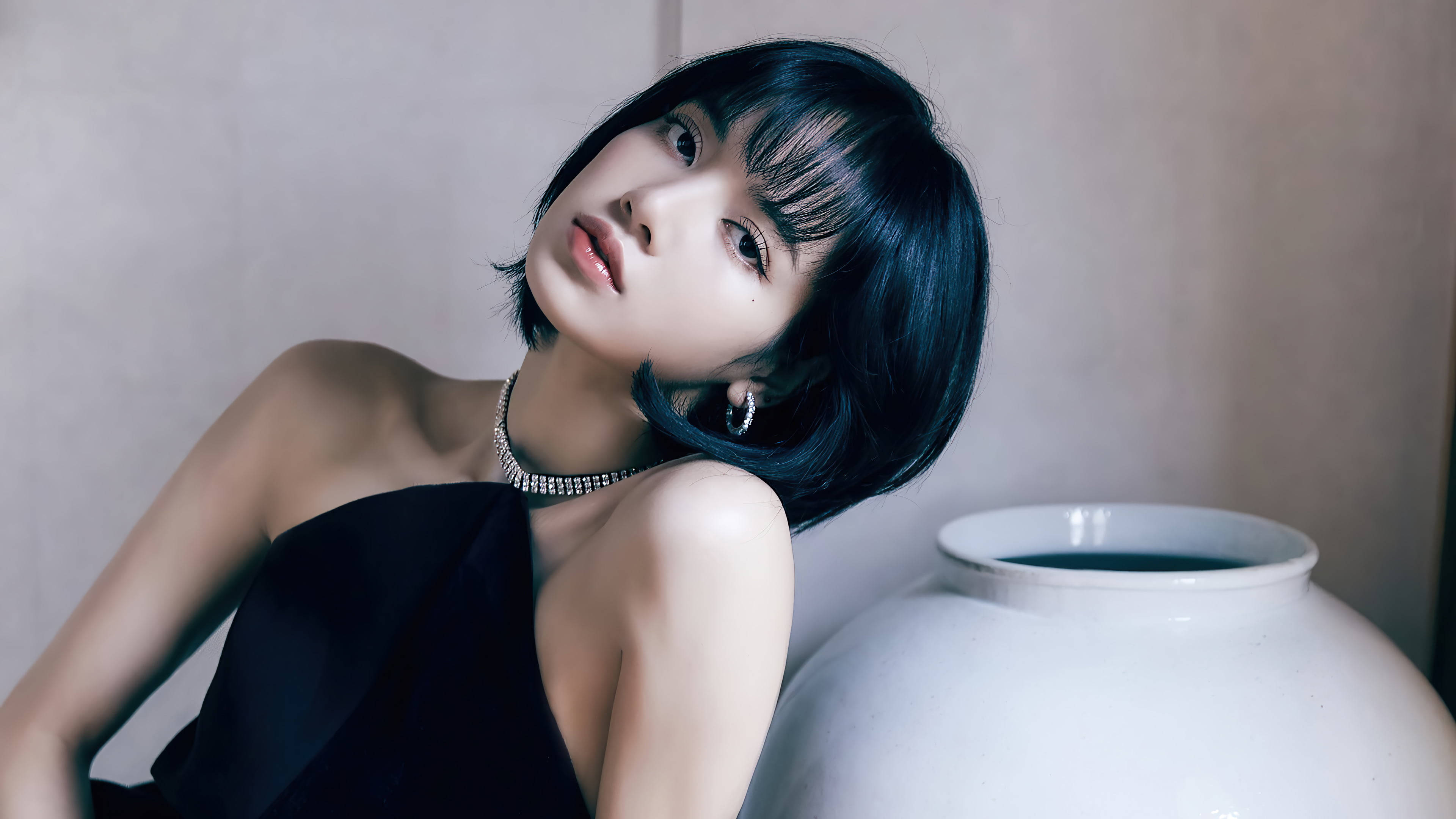 BLACKPINK 桌布 壁紙 — 一位短黑髮女性穿著黑色連衣裙和珍珠項鍊，在柔和的工作室燈光下靠著一個白色花瓶。