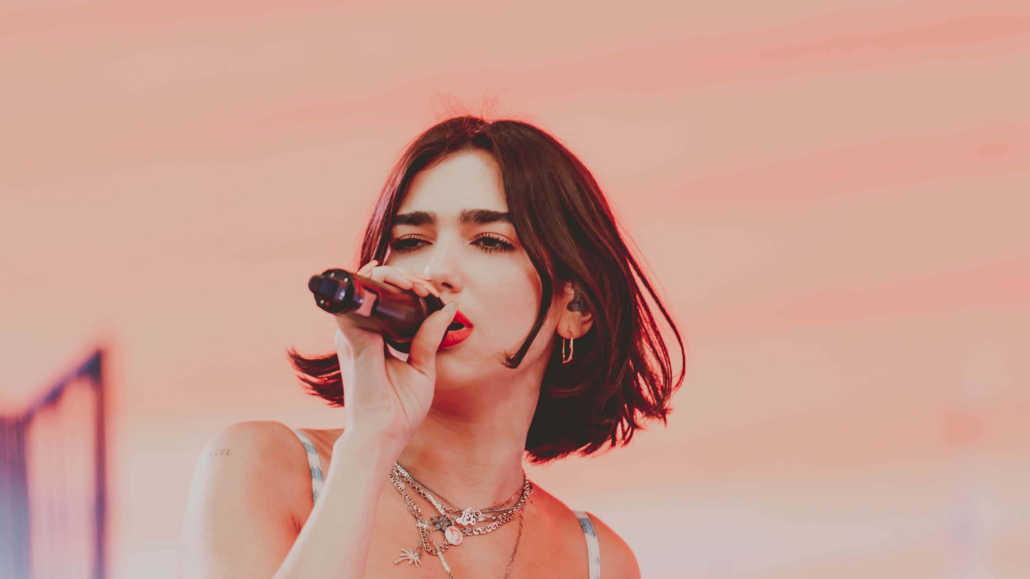 Dua Lipa 桌布 壁紙 — 一位短黑髮女歌手在柔和的粉紅和橙色漸變背景前，熱情地對著麥克風演唱。