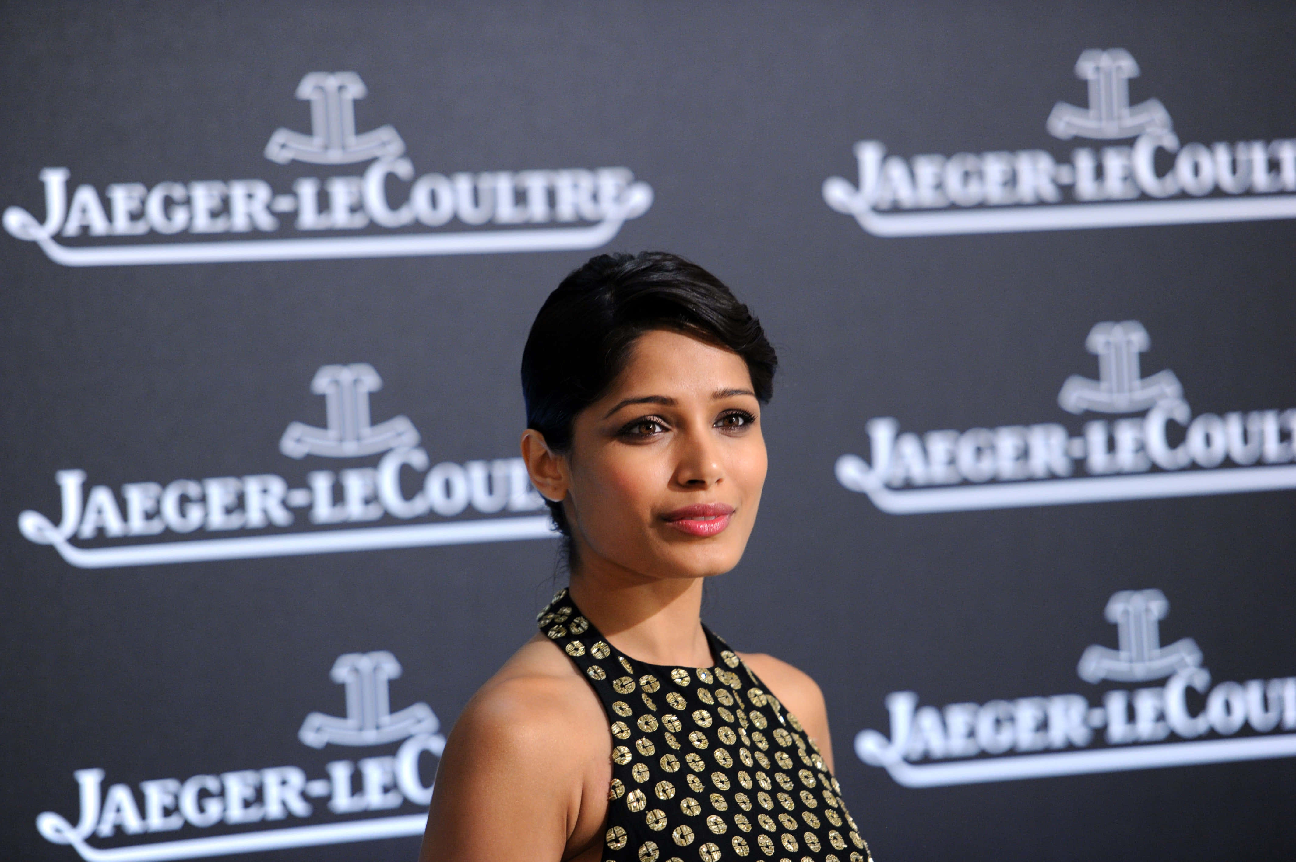 Freida Pinto 桌布 壁紙 — 一位短黑髮的優雅女性，穿著帶有金色圖案的黑色連衣裙，背景是重複的Jaeger-LeCoultre標誌。