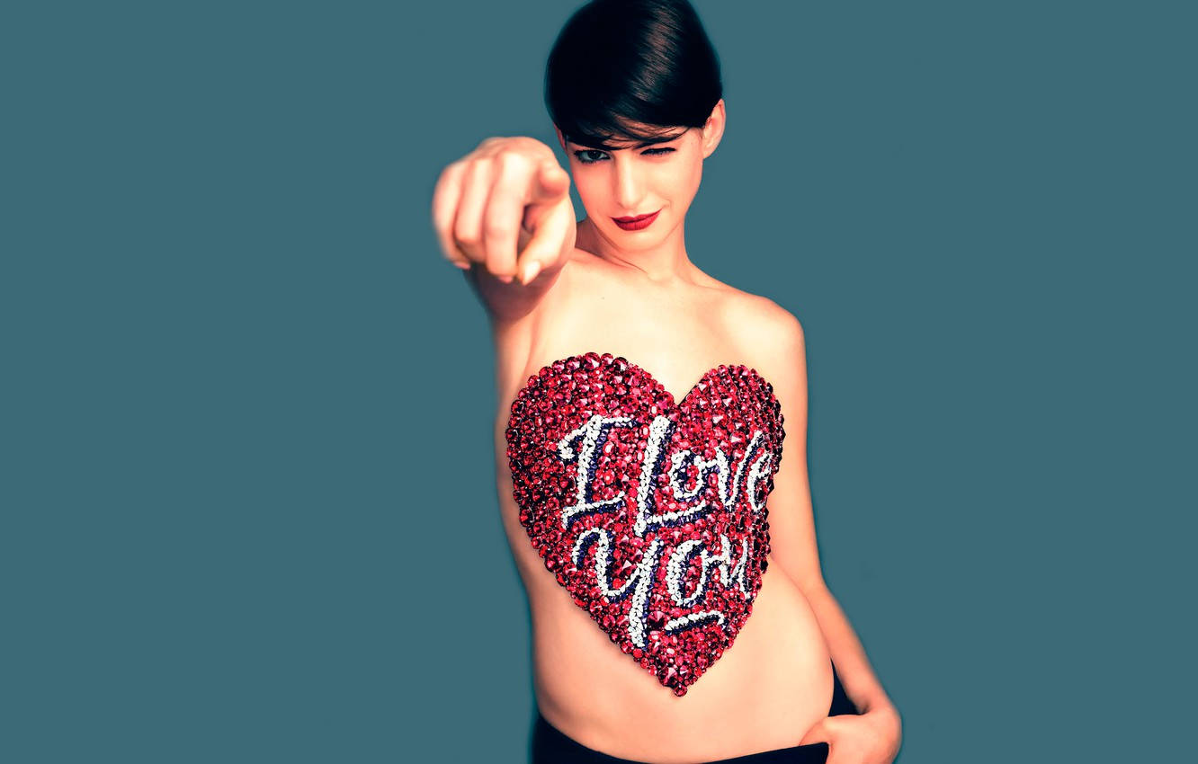 Anne Hathaway 桌布 壁紙 — 一位短黑髮的女性直接指向觀眾，胸前有一個閃閃發光的紅色心形，上面有白色字體寫著「I Love You」，背景為青綠色。