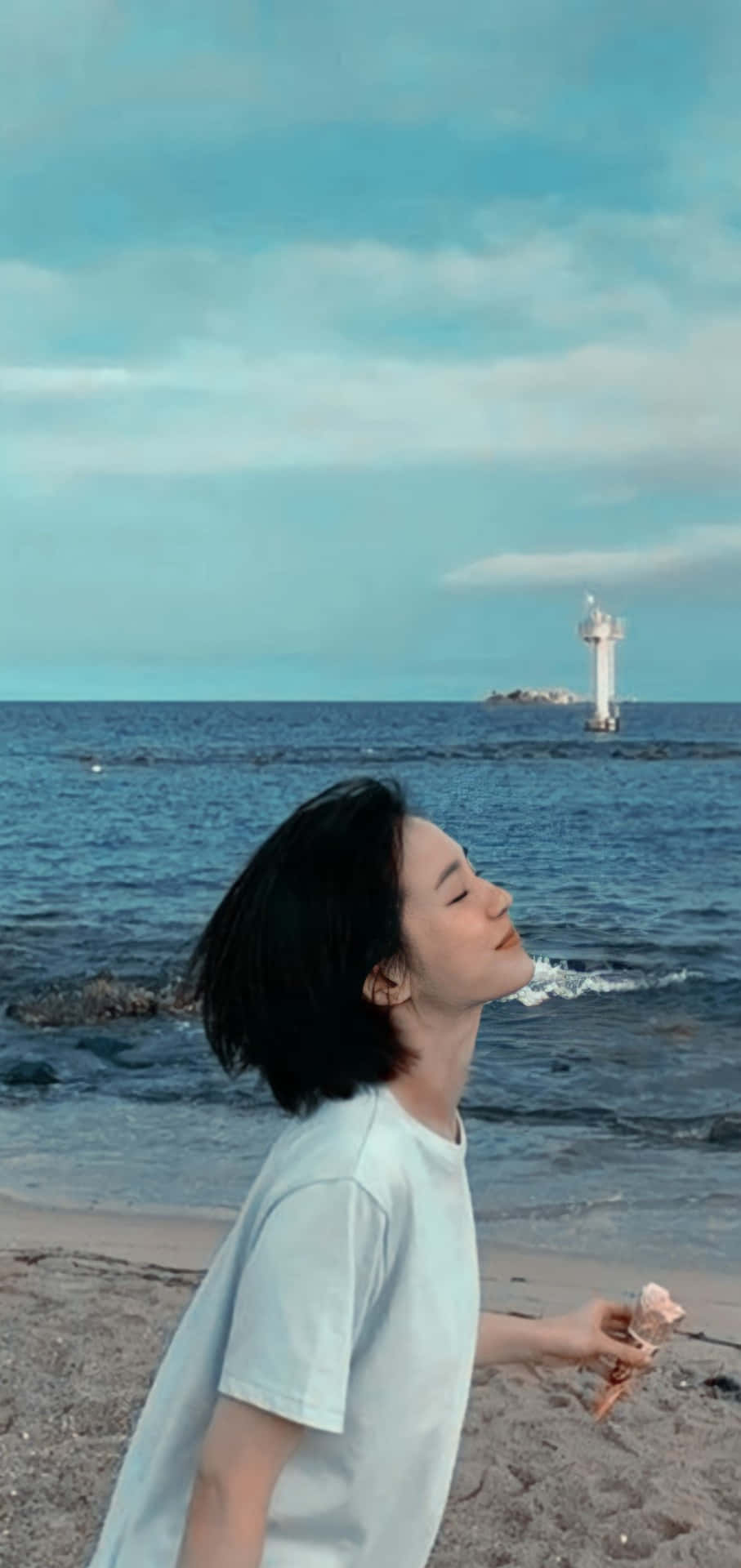 Park Gyu Young 桌布 壁紙 — 一位短黑髮的年輕女子站在岩石海灘上，閉著眼睛，享受著冰淇淋甜筒，遠處的燈塔在柔和的藍天下閃耀。
