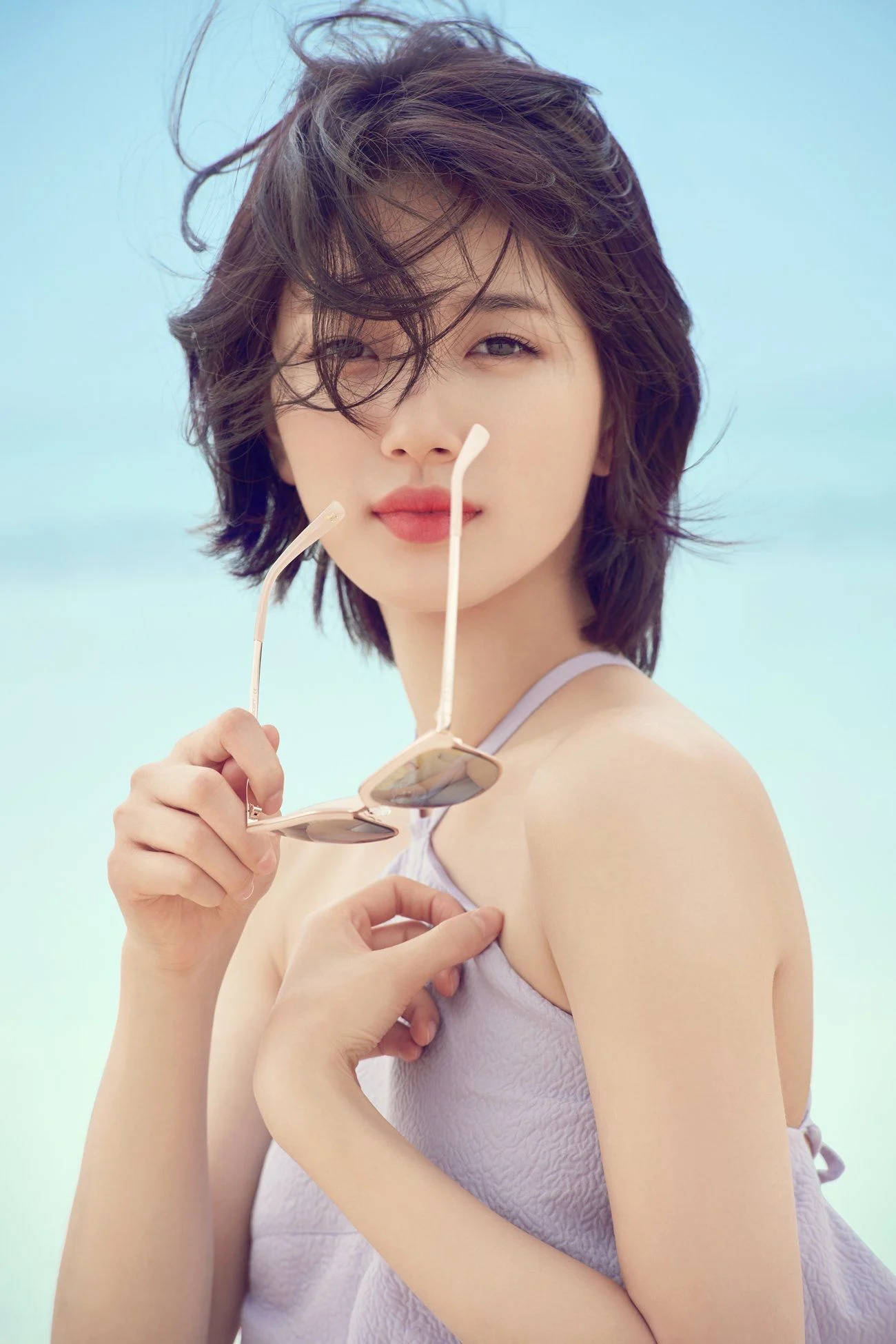 bae 桌布 壁紙 — 一位短黑髮的年輕女性，在柔和的藍天背景下將太陽眼鏡貼在唇邊。