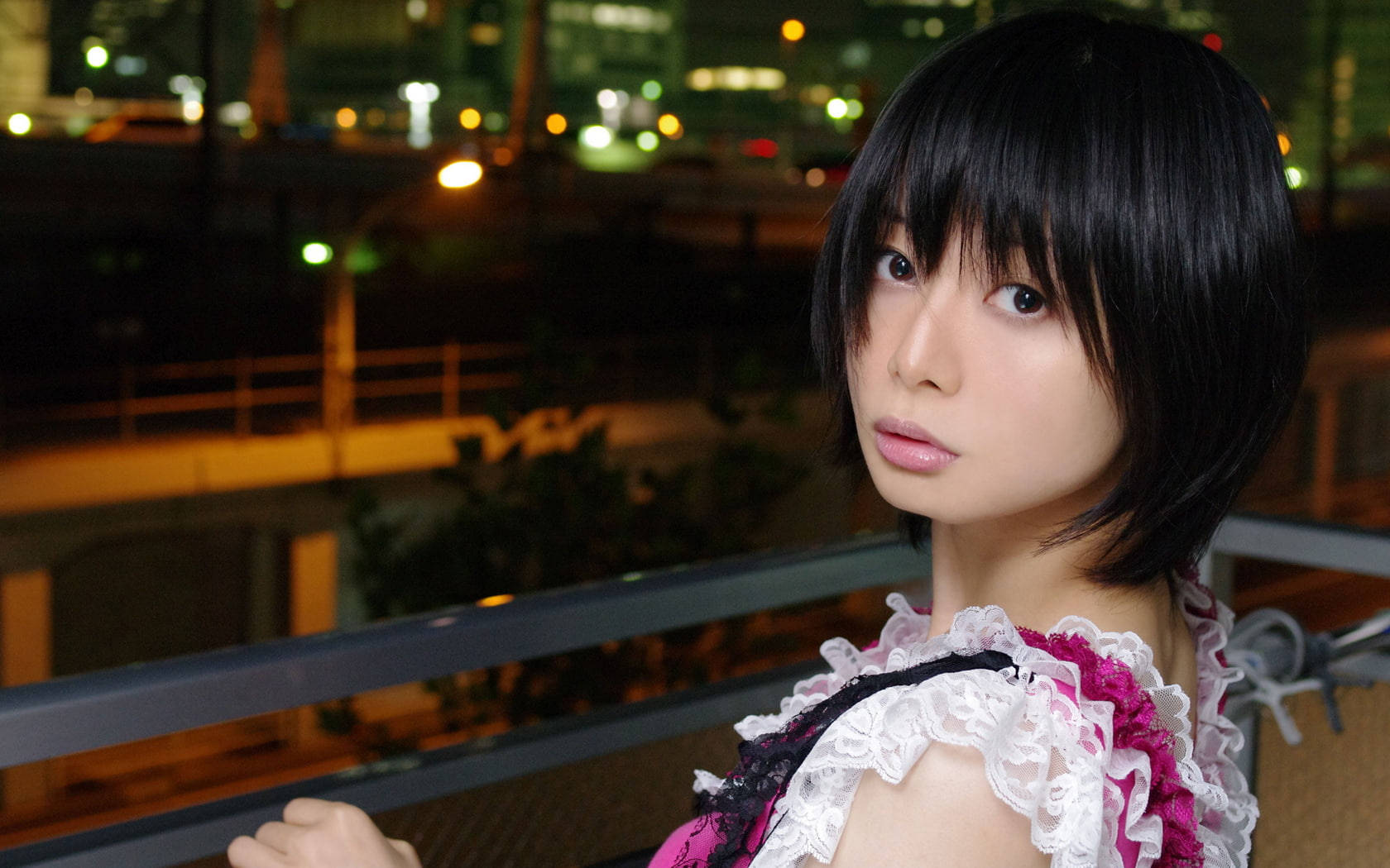 Cute Girls 桌布 壁紙 — 一位短黑髮的年輕女性，在模糊的城市夜景和閃爍燈光背景下，溫柔地凝視著鏡頭。