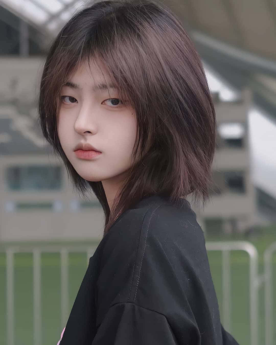 Hairstyle 桌布 壁紙 — 一位短黑髮的年輕女性，穿著黑色上衣，在模糊的體育場背景下微微轉頭。