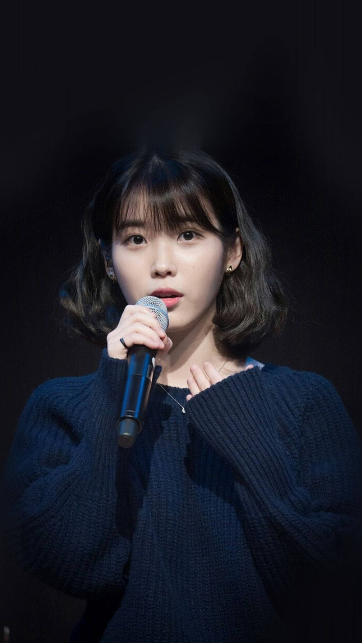 iu phone 桌布 壁紙 — 一位短黑髮的年輕女性在黑色背景前穿著海軍藍針織毛衣手持麥克風。