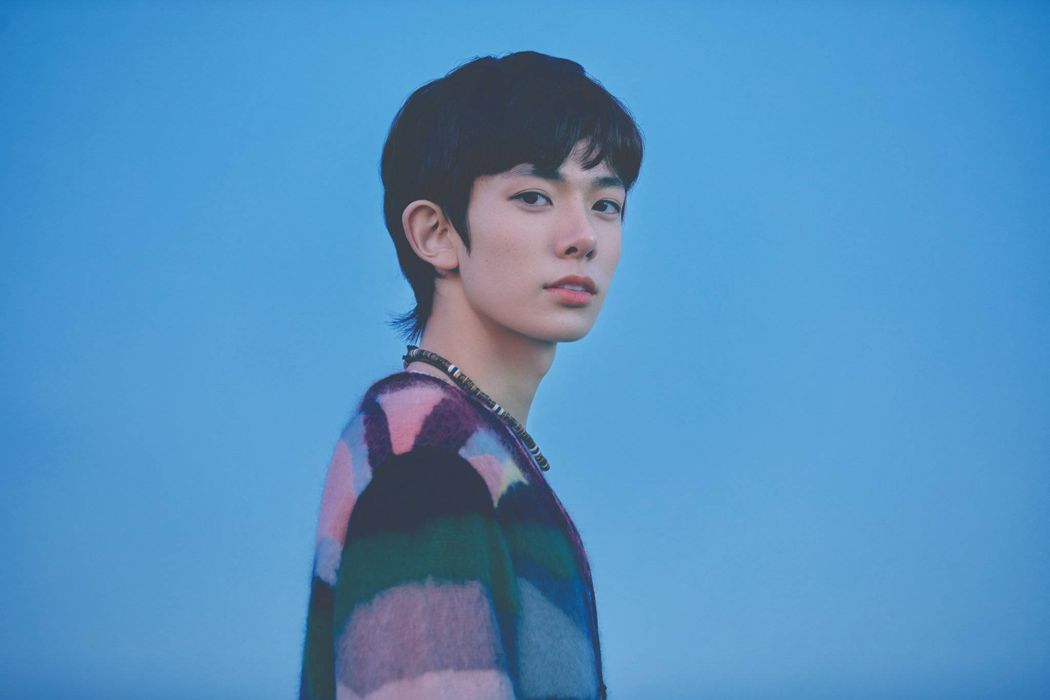 Enhypen Aesthetic 桌布 壁紙 — 一位短黑髮的年輕男子穿著色彩繽紛的拼布毛衣，背景為純藍色。