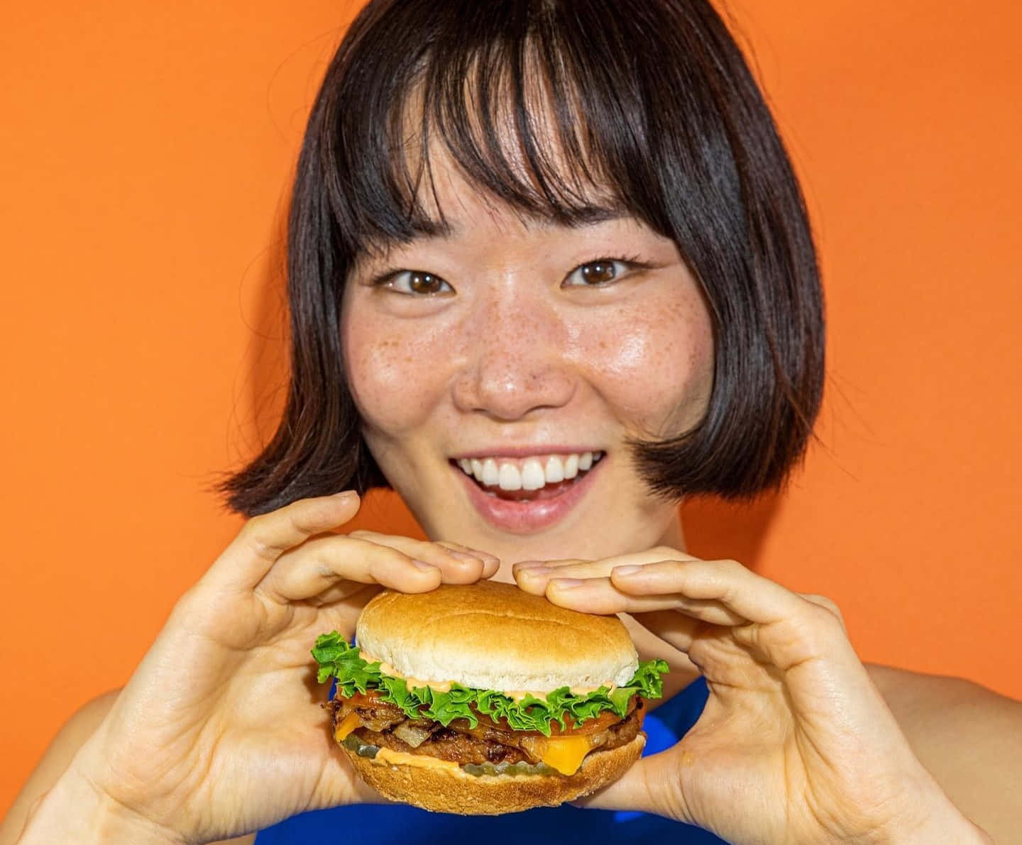 fast food 桌布 壁紙 — 一位短黑髮的快樂女性，在鮮豔的橙色背景前拿著多汁的起司漢堡，笑容燦爛。
