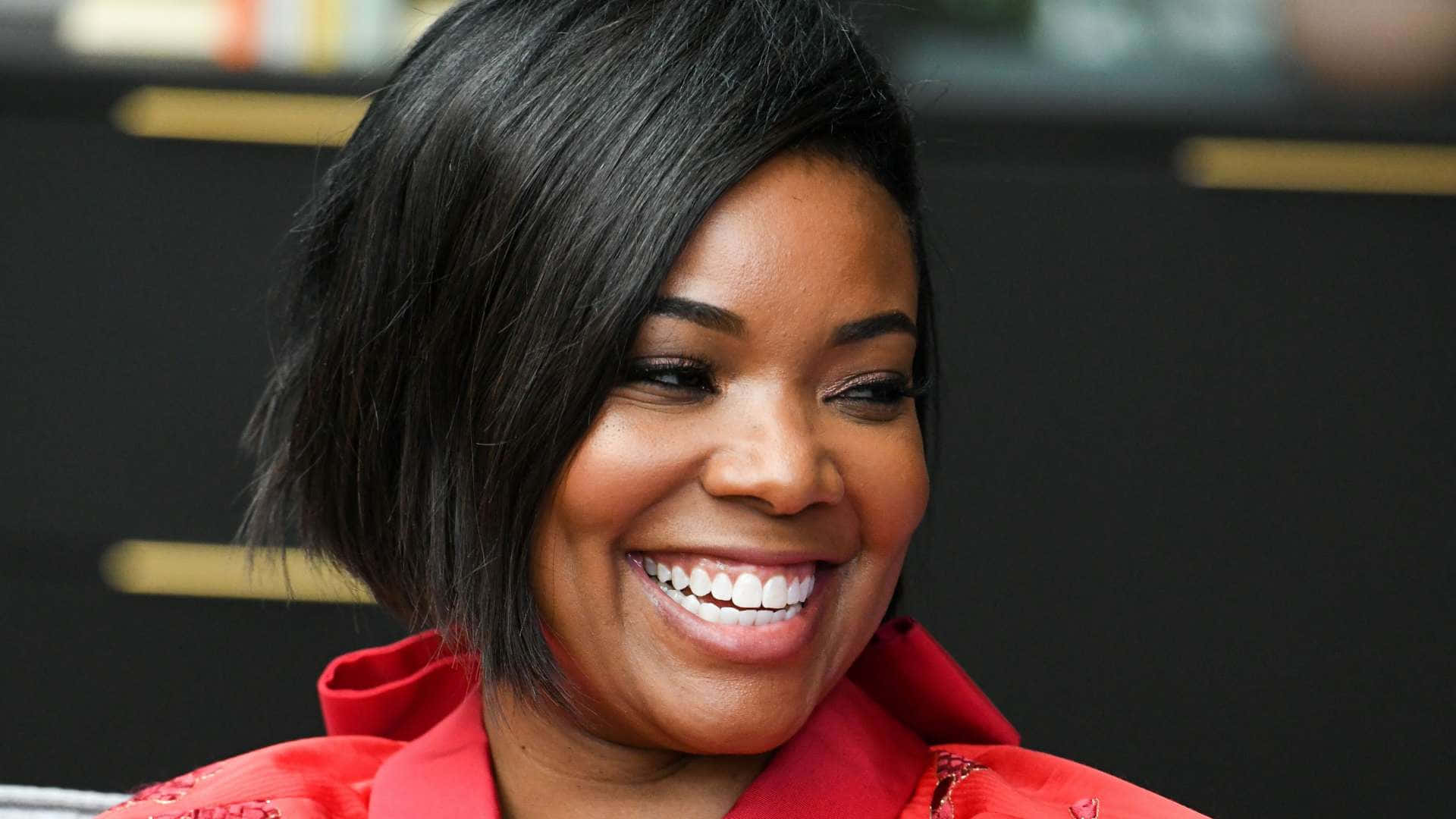 gabrielle union 桌布 壁紙 — 一位短黑髮的快樂女性，在黑暗模糊的背景中穿著鮮紅色外套，光彩照人。