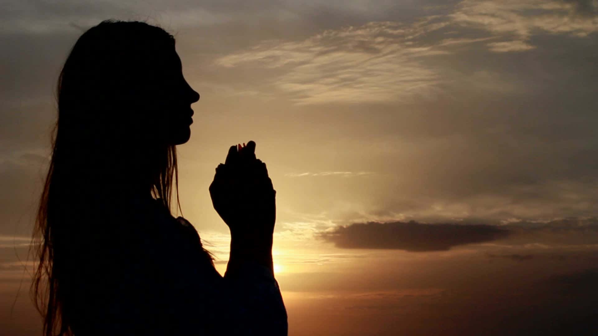 攝影 桌布 壁紙 — 一位祈禱中的女性剪影，背景是溫暖的日落天空和柔軟的雲朵。