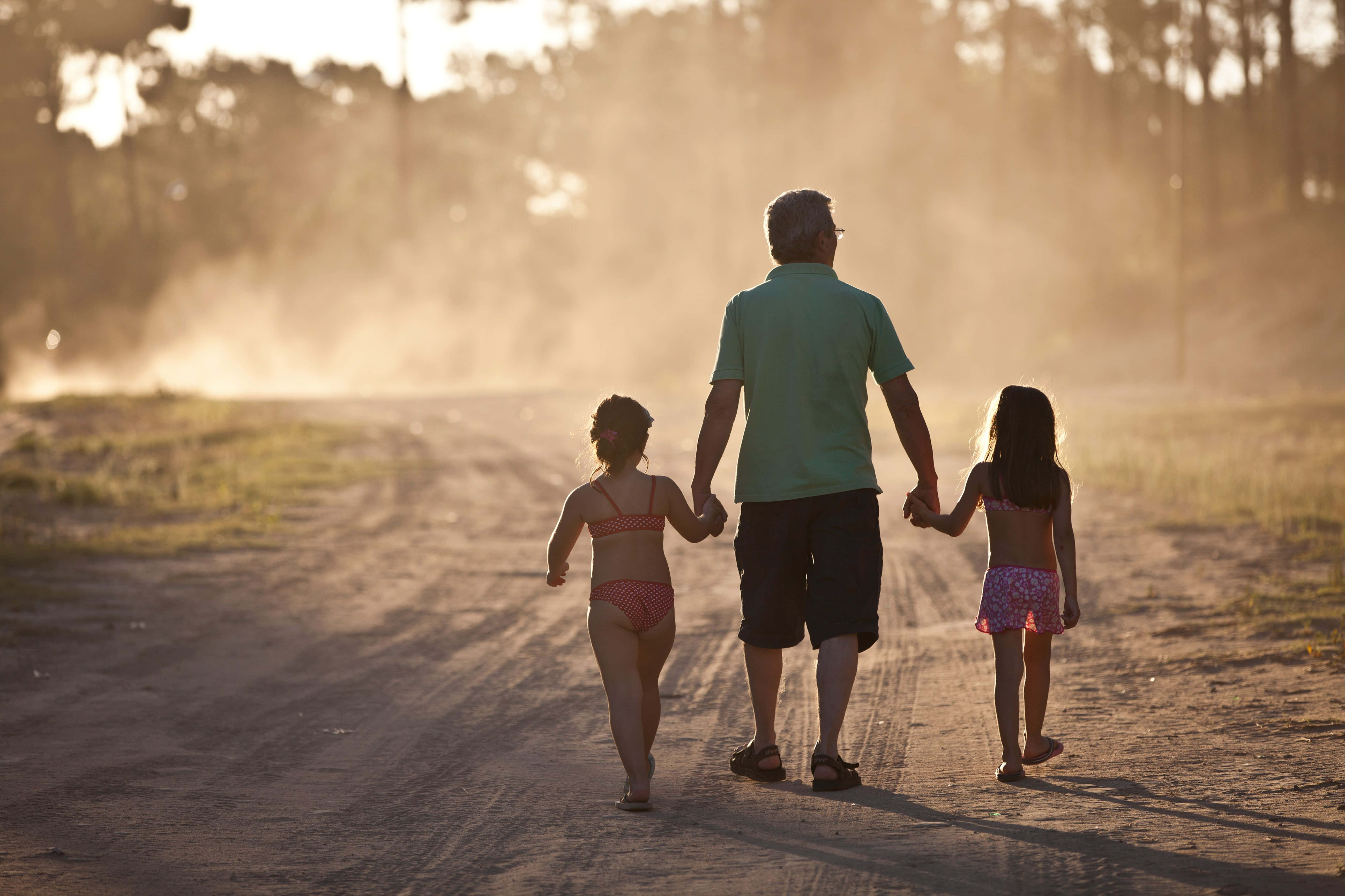 Family 桌布 壁紙 — 一位祖父與兩個小女孩手牽手，走在沐浴在金色夕陽光下的塵土飛揚鄉村小路上。