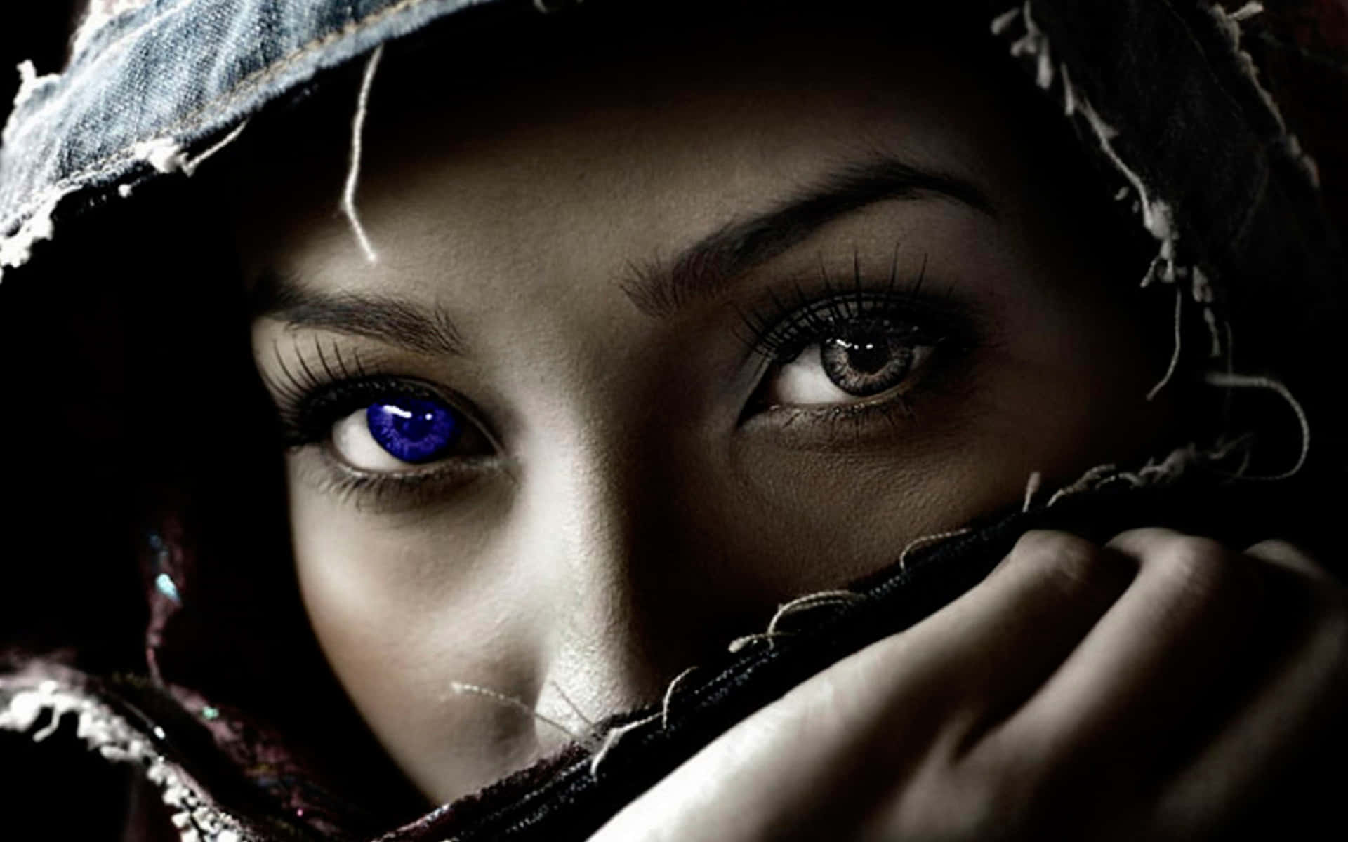 Eye 桌布 壁紙 — 一位神秘女子，一隻眼睛發出藍光，另一隻眼睛是棕色，透過破爛的兜帽在陰暗的黑暗中凝視。