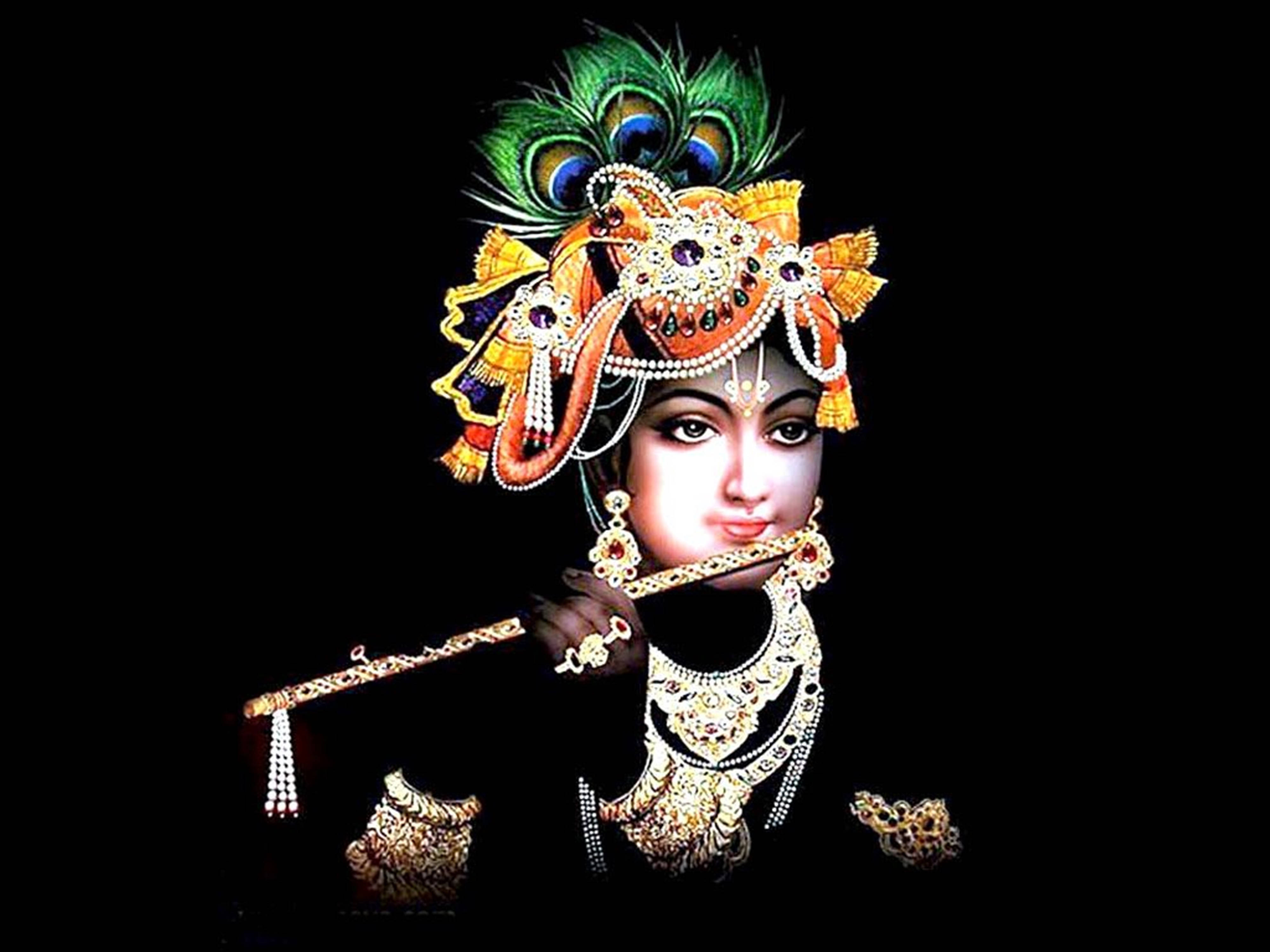 Krishna Flute 桌布 壁紙 — 一位神聖人物佩戴金色珠寶和孔雀羽毛冠冕，在純黑背景前吹奏長笛。
