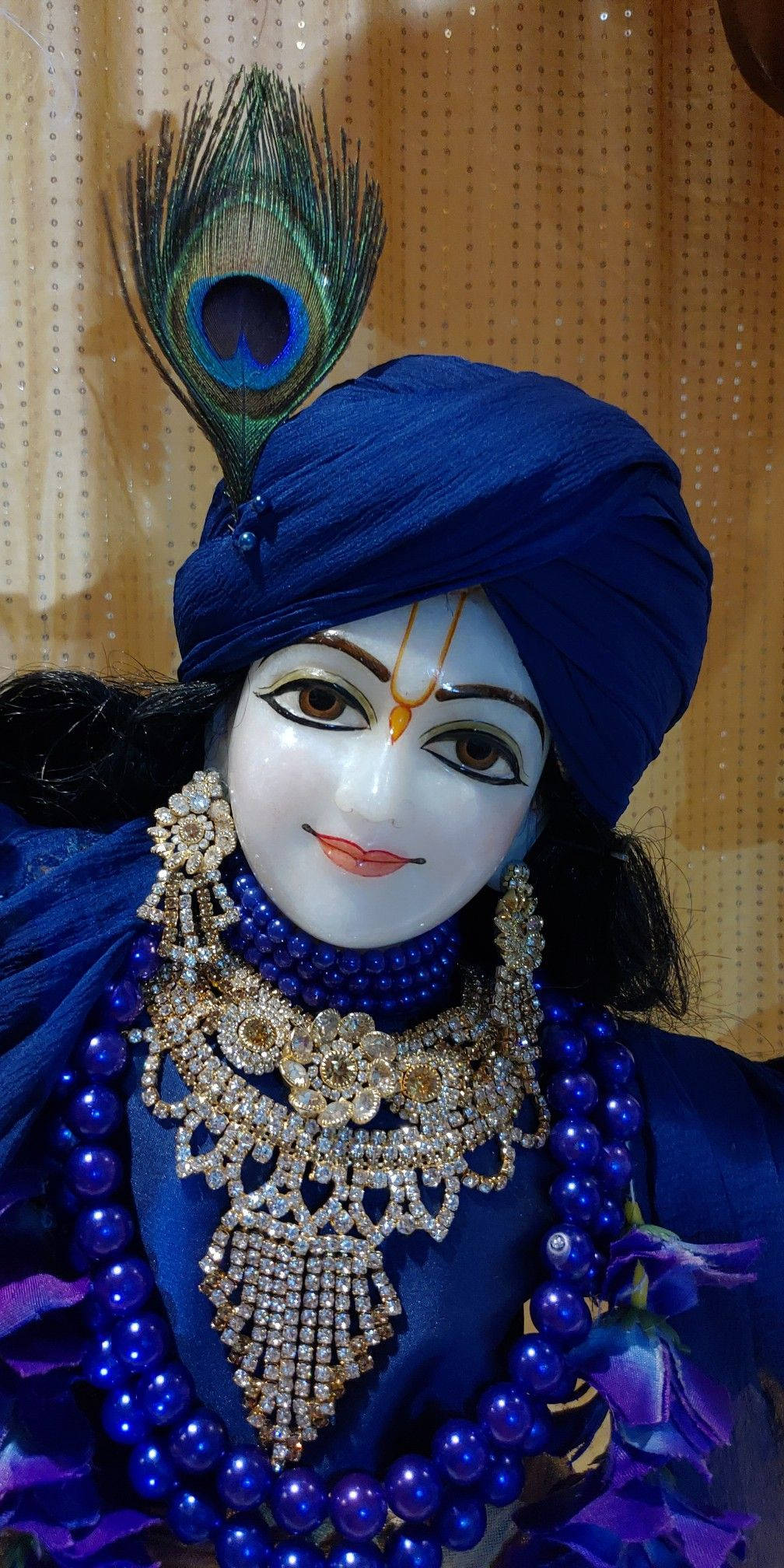 krishna phone 桌布 壁紙 — 一位神聖人物身穿皇家藍色服飾和珠寶，頭戴孔雀羽毛冠冕，背景是點點窗簾。